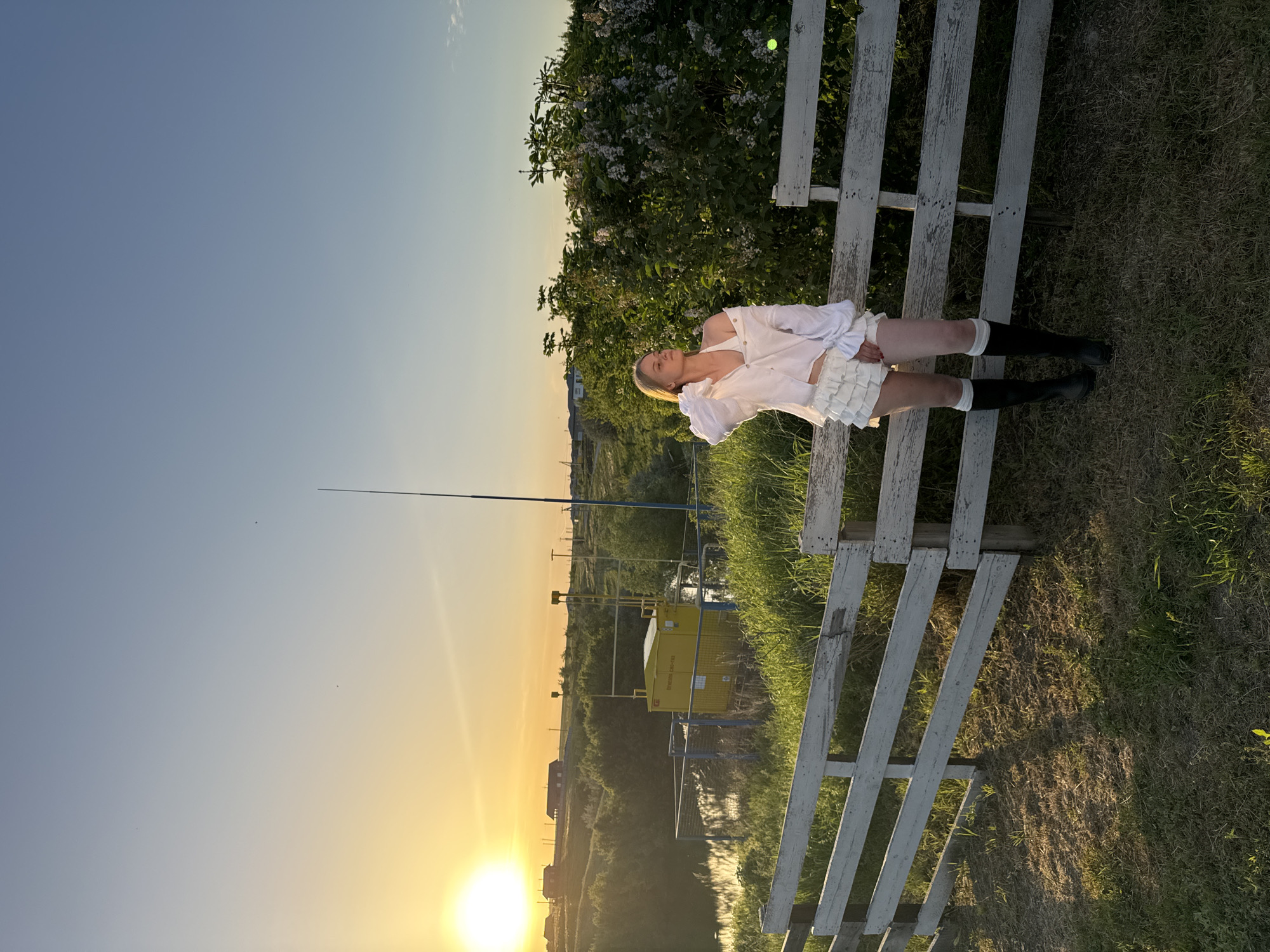 The image shows a woman lying on a white wooden fence. She is wearing a white dress and black boots with white socks. Behind her is a lilac bush. In the background, there is a rural landscape with a sunset and a gas station. The scene is located in Усть-Курдюм, Russia.