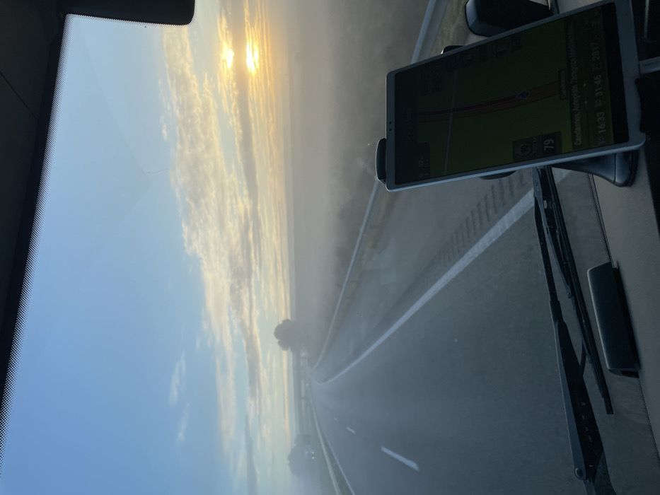 The image presents a first-person view from inside a vehicle, looking out through the windshield onto a multi-lane highway. The scene is enveloped in dense mist or fog, which heavily obscures the distant road and surrounding landscape, though faint outlines of trees are visible along the roadside. The sky above, seen through the windshield, is a soft blue, with a low sun casting a golden glow through scattered clouds and the pervasive atmospheric haze. This lighting suggests a late afternoon setting, consistent with the time displayed on the navigation device.
A tablet-like navigation device is mounted on the right side of the windshield, displaying a map and route. Several pieces of text are clearly visible on its screen: "Candasnos, Autovía Zaragoza Tarragona Mediterráneo," which specifies the location in Spain. The device also shows a current speed of "79" (likely km/h), alongside speed limit indicators of "80" and "100." Additional information includes a remaining distance of "281.7km" and a precise time of "16:33."
Parts of the vehicle's windshield wiper blades are visible in the lower right corner of the frame. A minor, faint crack or imperfection is discernible on the upper left portion of the windshield. The overall atmosphere evokes a sense of serene, yet cautious, travel through the misty late afternoon conditions on the highway.