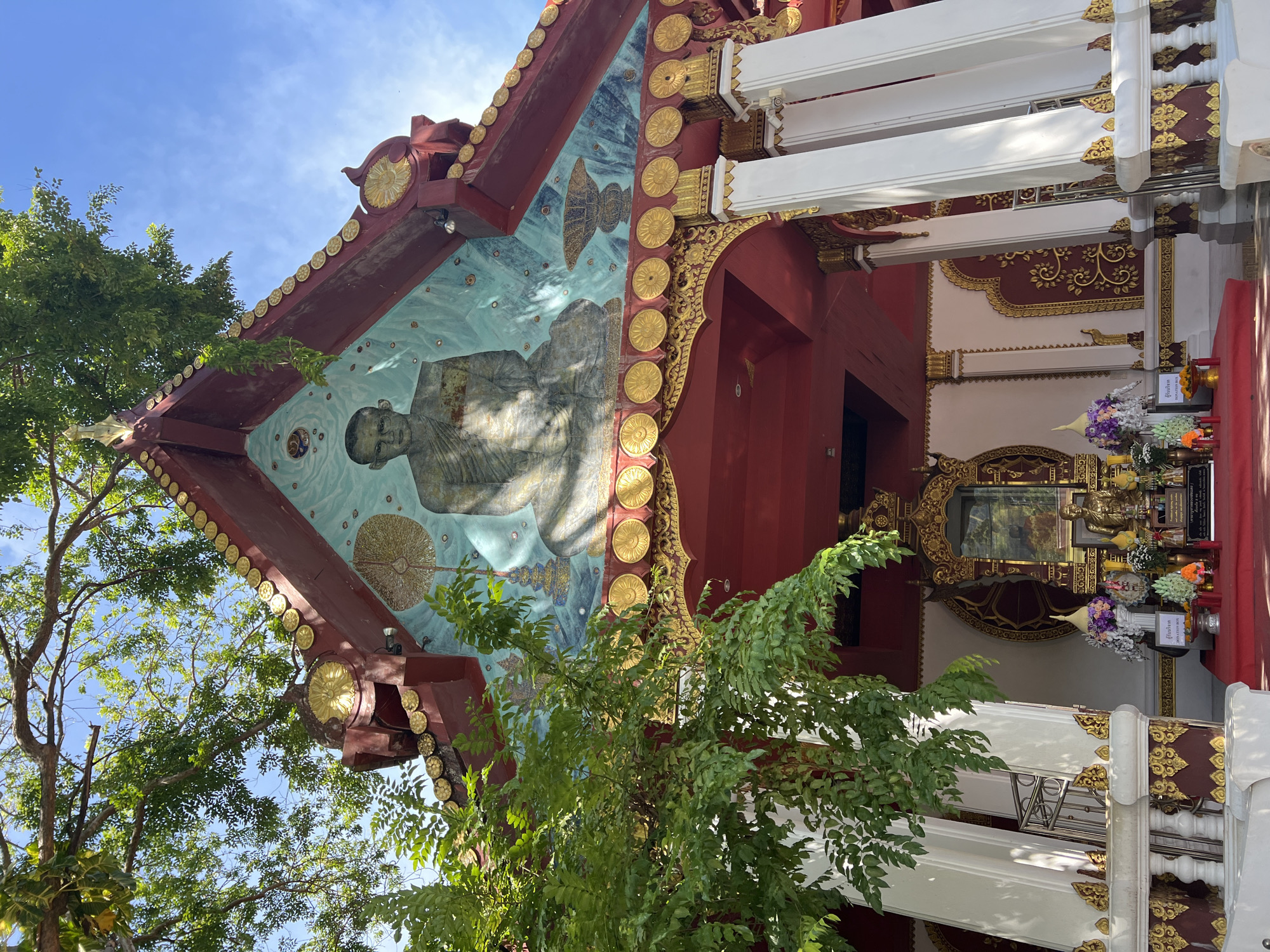 A low-angle outdoor shot captures the ornate facade of a temple structure in Baan Hua Thanon, Thailand, under a clear blue sky. Lush green foliage from a large tree partially obscures the upper left portion of the temple. The temple's pediment is a vibrant mural featuring a serene portrait of a seated monk, rendered in blues, grays, and yellows, surrounded by abstract circular motifs and a tiered pagoda. The temple's roofline is decorated with rows of gold, coin-like embellishments and a deep red trim. White pillars with gold accents support the structure, leading to an entrance where a red carpet leads to an altar. The altar is adorned with a statue, floral arrangements, and offerings. The overall scene suggests a place of worship, characterized by rich cultural and religious artistry.