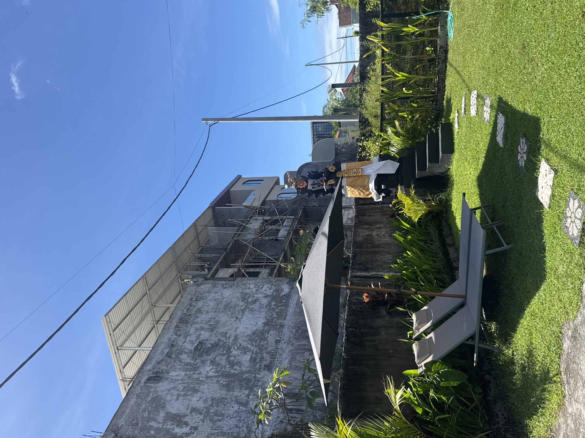 The image shows a partially constructed building in Kuta Selatan, Indonesia. The building has a concrete exterior with exposed scaffolding. An electrical pole is visible in front of the building. In the foreground, there is a garden with plants, grass, stepping stones, and two gray lounge chairs. There is a structure with a yellow cloth draped over it and several people behind it, close to the fence. The sky is blue and cloudless. A partially open umbrella stands between the camera and the building.