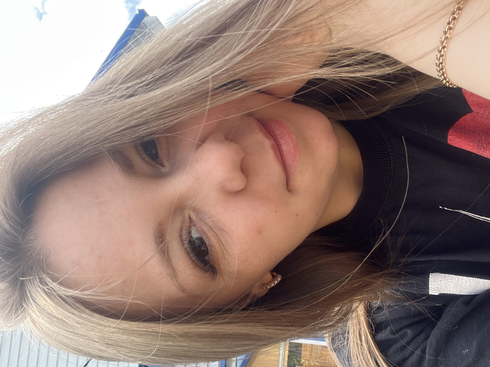 The image is a close-up, selfie-style photograph of a young Caucasian woman. She has fair skin, blonde hair, and is looking towards the camera with a slight smile. Her right eye is more prominently visible, showing dark irises and long eyelashes. She is wearing a dark crewneck shirt. A hint of a building with blue accents is visible in the background, as is a bright sky with a few clouds, suggesting an outdoor setting. The woman is wearing a small, sparkly hoop earring in her visible ear, and a delicate gold chain bracelet on her wrist. The scene does not contain any identifiable landmarks or specific indicators that would allow for its location to be determined within the city of Shadrinsk, Russia.
