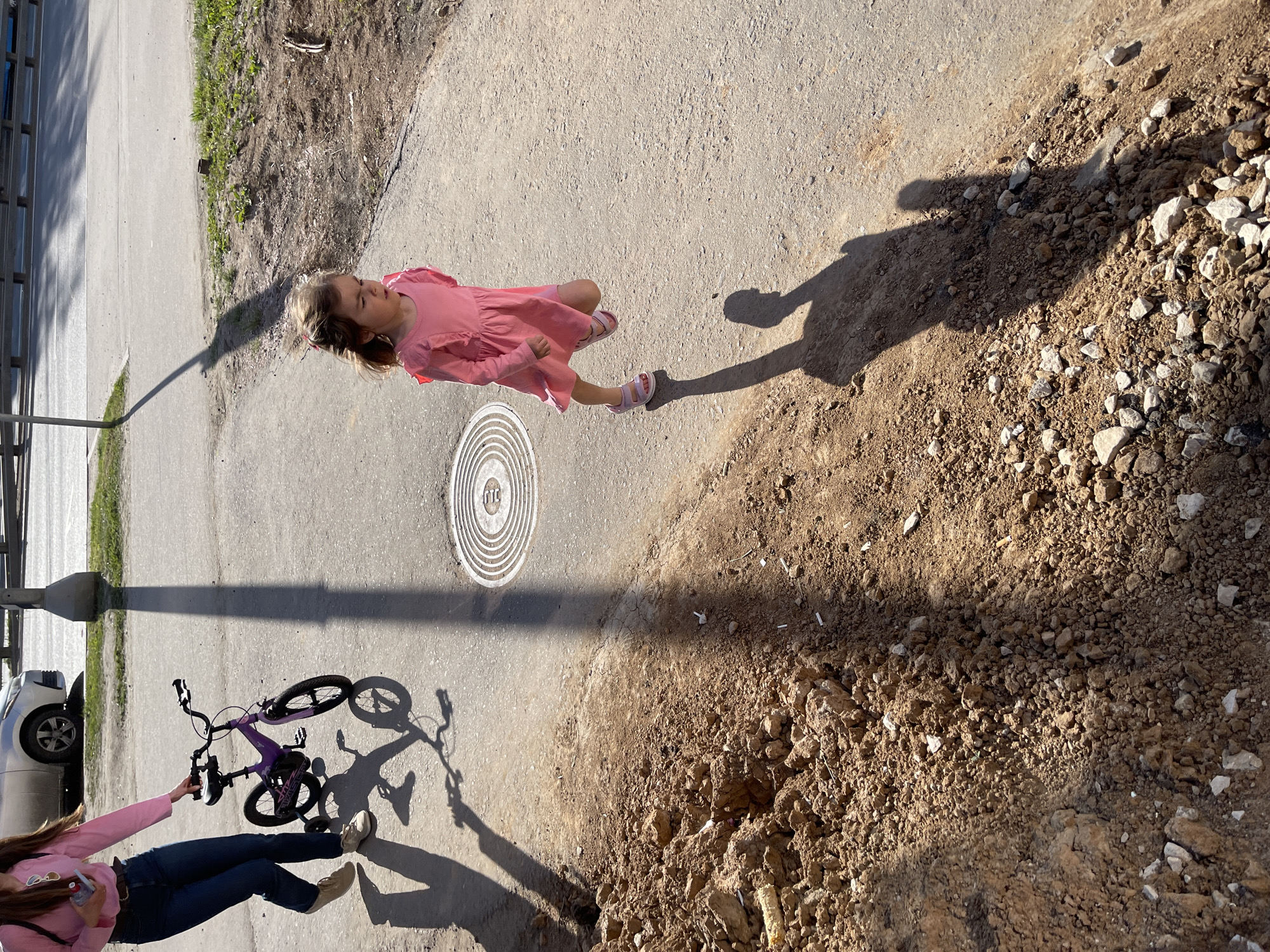The image captures an outdoor scene in Kommunarka, Russia, on a sunny day with distinct long shadows. The perspective is an overhead shot looking down at the ground and subjects.
The main subject is a young child, likely a girl, with light brown hair, wearing a pink ruffled dress and pink sandals. She stands on a paved concrete or asphalt path, looking slightly upwards and to her right. Her elongated shadow stretches out ahead of her onto a large pile of brown dirt and small, light-colored rocks and gravel that flanks the path.
In the lower-left portion of the image, an adult (partially visible) is present, wearing a pink long-sleeved top, blue jeans, and light-colored sneakers. This person is holding a purple child's bicycle, which has training wheels. The adult and bicycle also cast long shadows across the paved surface. A silver car is partially visible parked behind them.
The ground consists of a wide paved pathway, a narrow strip of patchy grass and soil, and the prominent mound of loose earth and gravel. Embedded in the paved path is a circular manhole cover with a pattern of concentric rings. The letters "МТС" are clearly visible at the top of the manhole cover, indicating a connection to the Russian telecommunications company.
The strong, elongated shadows suggest the time of day is either early morning or late afternoon. The scene depicts a casual outdoor activity, possibly a stroll or a break from cycling, in what appears to be a suburban or a developing area with a mix of paved surfaces and natural, uncultivated ground. No specific emotions are discernible from the child's expression.