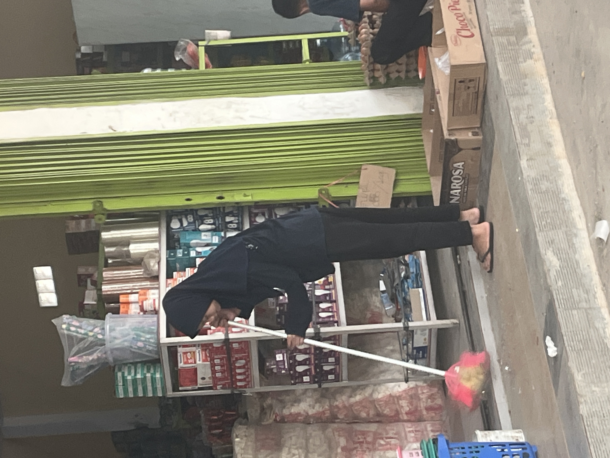 In Sekayu, Indonesia, a person wearing a black hijab and dark clothing is sweeping the ground in front of a store with a green metal grate. The person is holding a broom with a pink and yellow brush. The store appears to sell various goods, with shelves stocked with boxes and containers visible in the background. A blue plastic basket is on the left side of the image. Further to the right, two cardboard boxes are on the ground, one labeled "NAROSA" and the other "Choco Pie". A person in dark clothing is partially visible on the far right, sitting down and facing away from the camera. The ground is paved with concrete steps.