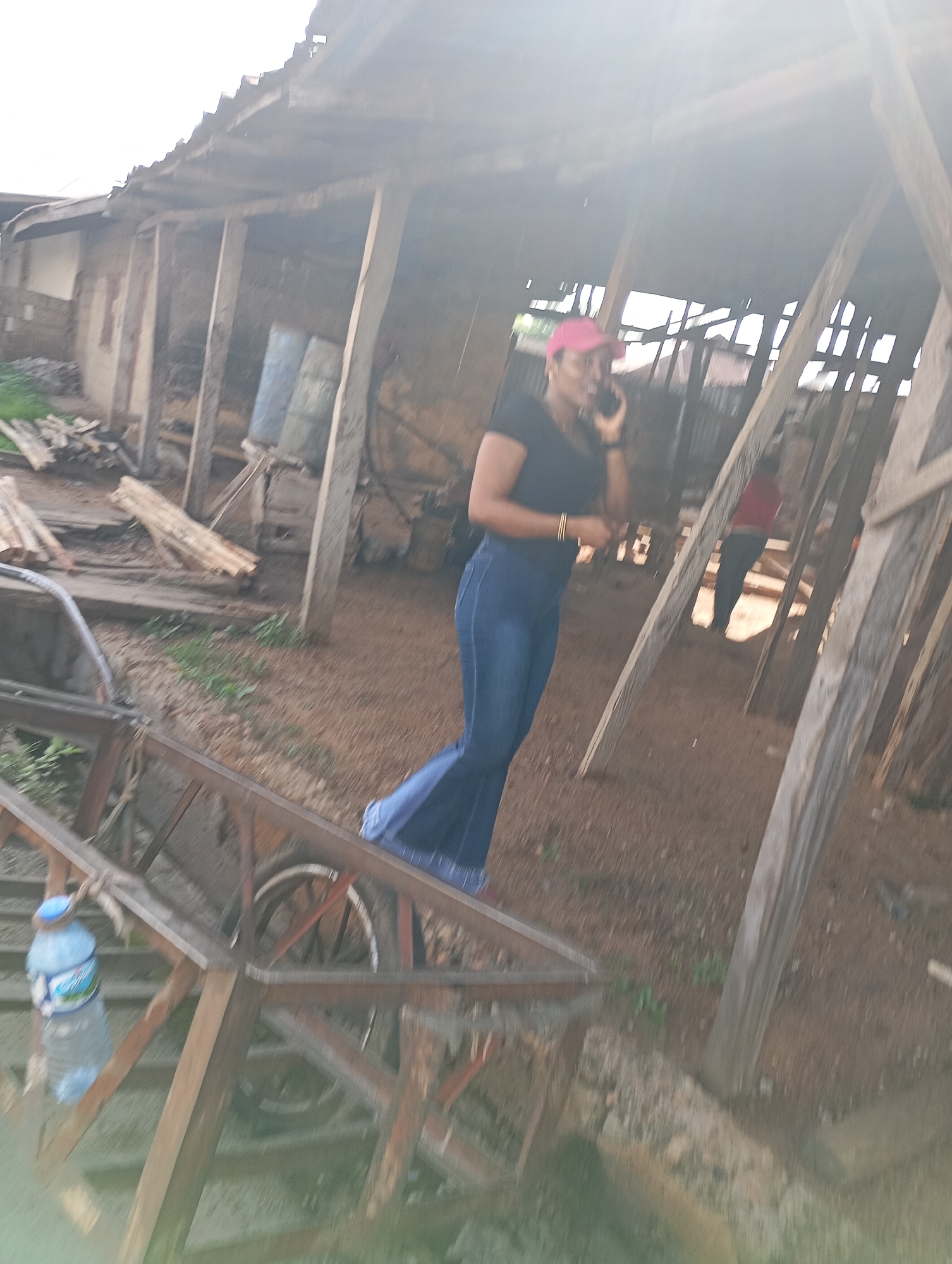 A woman wearing a pink cap, black shirt, and blue jeans is standing on a construction site in Shendam, Nigeria, talking on her phone. She is the main subject of the image and is positioned in the center-right. The construction site appears to be outdoors with a partially constructed wooden structure. There are other people and objects in the background, including lumber, a water bottle, and a wheelbarrow. The overall scene is blurry due to motion, suggesting the photo was taken with a moving camera or in low light conditions. The ground is covered with dirt and debris. The sky is overcast and bright.
