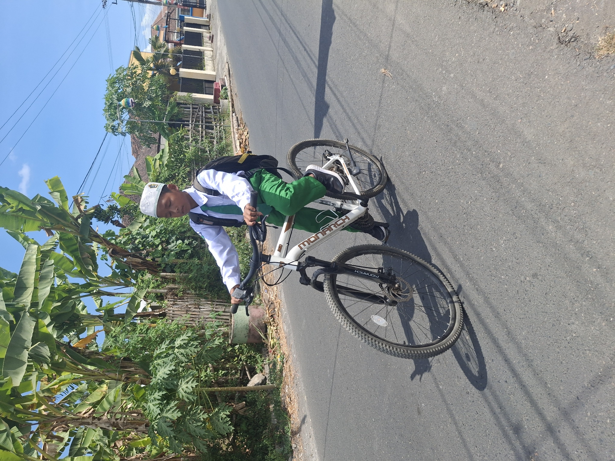 In Denok, Indonesia, a young boy dressed in a white school uniform with a green tie and a white peci (a cap) rides a white bicycle. He wears green pants and a backpack. The boy is on a paved road, with lush green vegetation and trees lining the left side of the road. In the background, there are houses and a bright blue sky with some clouds. The sun casts shadows on the road, indicating it is daytime.