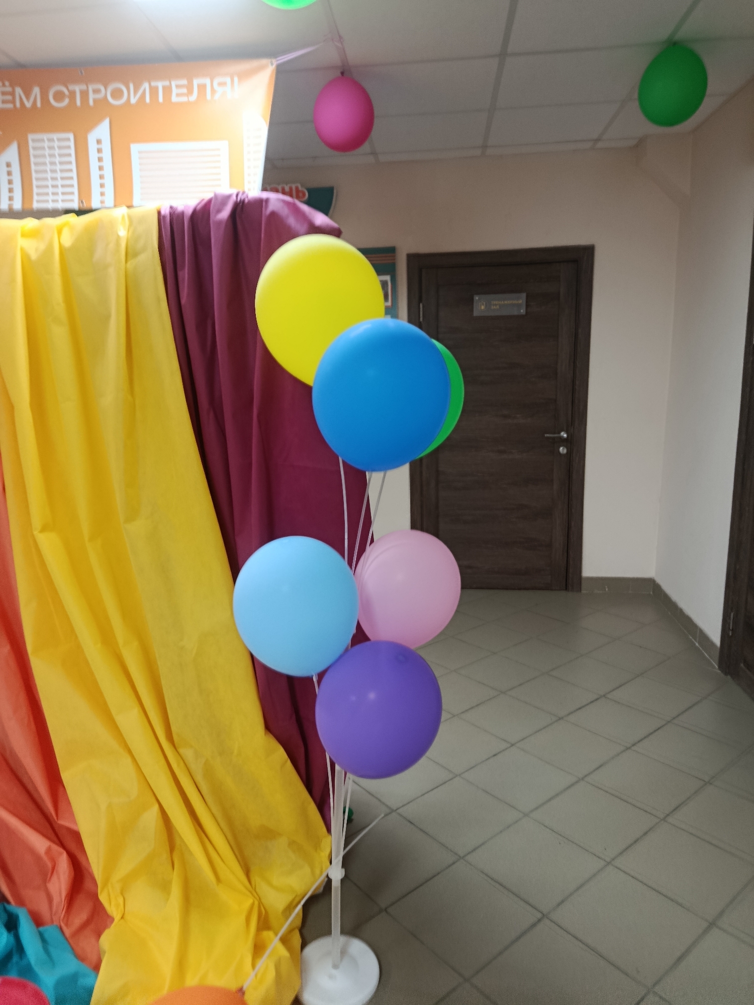 A cluster of five balloons on white stands is positioned in the foreground, adjacent to draped fabric in shades of yellow, burgundy, and orange. The balloons are colored yellow, blue, light blue, pink, and purple. In the background, two dark wood doors are visible on a neutral-colored wall, with one door slightly ajar. The floor is tiled in a light gray diamond pattern. A banner with white text and graphics is partially visible at the top, suggesting a celebratory or event-related setting. The scene appears to be indoors, possibly in a hallway or common area. There are no people or animals present in the image.