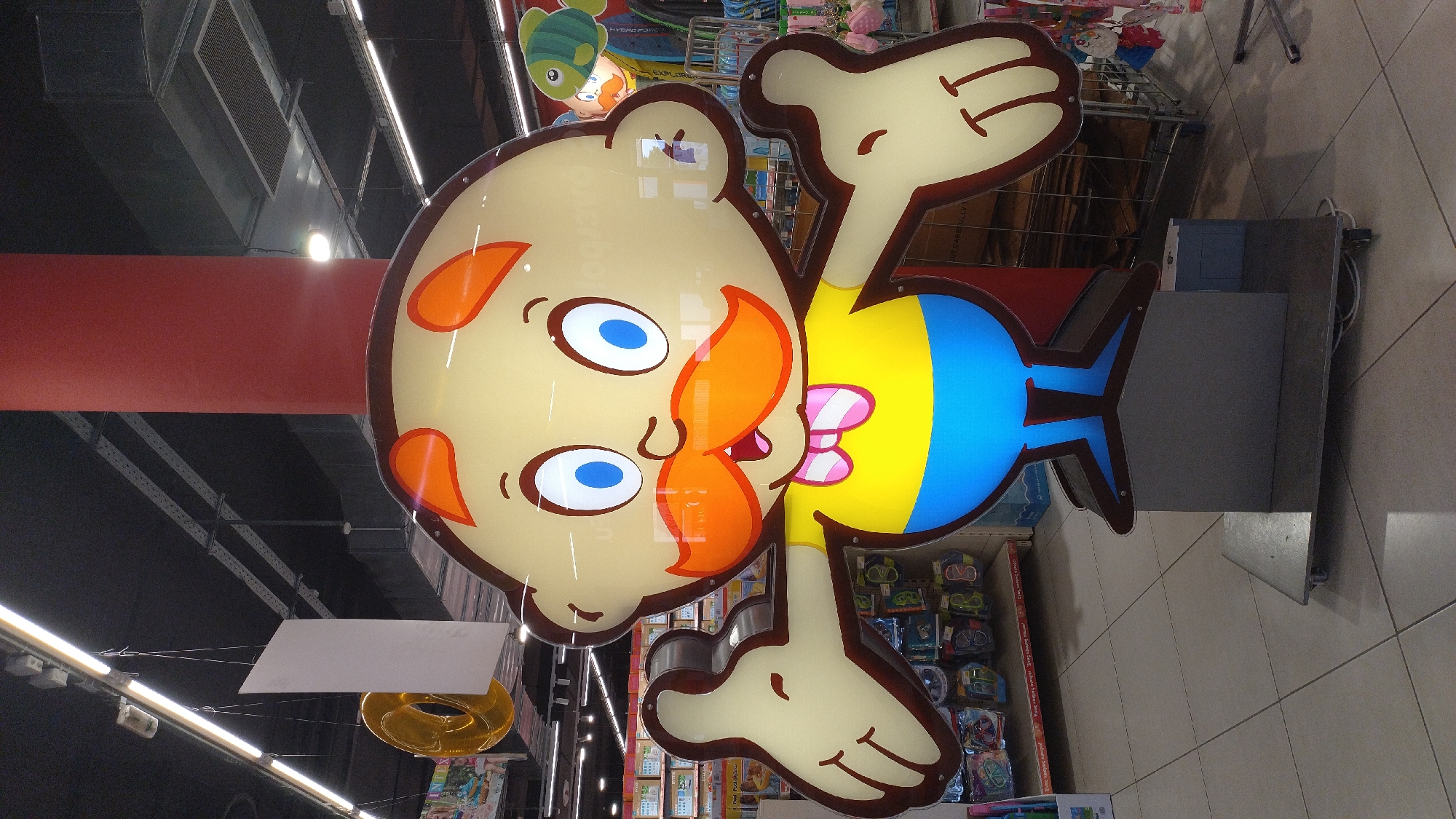 A large, illuminated, cartoon-like cutout of a man stands in the middle of a store. The man has a round, yellow head with orange eyebrows and ears. He has large blue eyes, a prominent orange mustache, and a pink and white striped bowtie. His body is yellow on top and blue on the bottom, resembling a shirt and pants. His arms are outstretched, and his hands are in a thumbs-up gesture. The cutout is mounted on a grey pedestal with wheels. In the background, shelves of products are visible, along with ceiling lights and a red pillar. The floor is tiled with a square pattern. The overall scene appears to be a retail environment, likely a toy store or a department store, within the city of Pylaia Municipal Unit, Greece, based on the provided information.