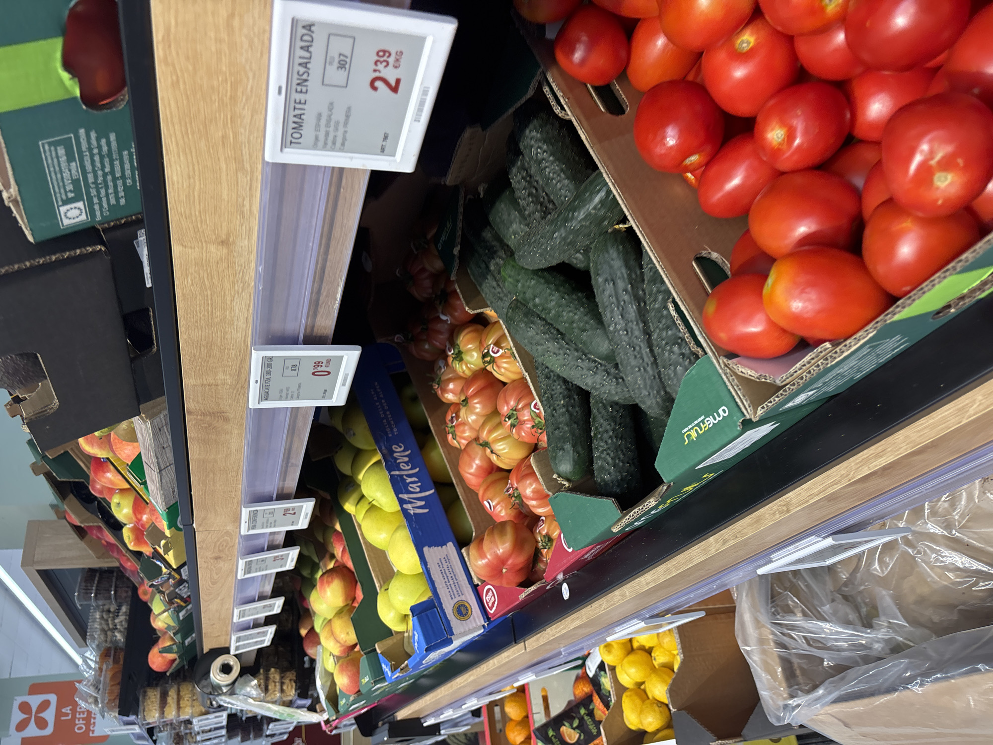 The image depicts a well-stocked produce aisle in a supermarket, likely located in Urbanización Villa Martín, Spain. The aisle features multiple shelves displaying a variety of fresh fruits and vegetables. In the foreground, boxes overflow with bright red tomatoes and dark green cucumbers. Further back, crates of apples and lemons are visible. The shelves are organized with clear price labels, one of which reads "TOMATE ENSALADA" with a price of 2.39 € per kilogram. The overall environment suggests a clean and organized retail space, typical of a Spanish grocery store. No people are visible in the frame.