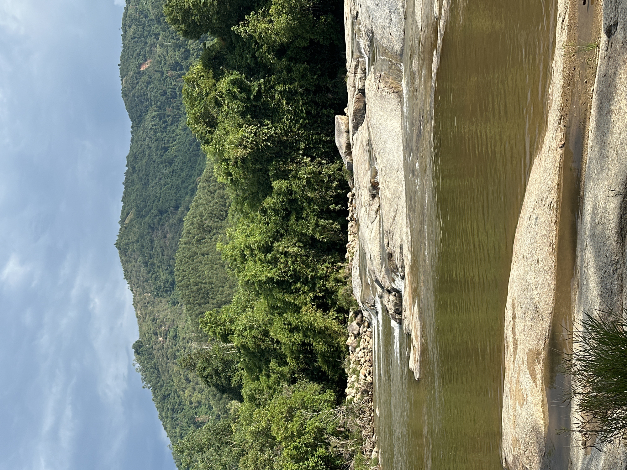 A wide shot captures a natural river scene in Suối Dầu, Vietnam. The foreground features a large, flat, light-colored rock formation with a small pool of water. The river, a muted olive green, flows from right to left, with ripples visible on its surface. The middle ground shows a series of smaller, uneven rocks and a gentle cascade of water. Behind the river and rocks, a dense wall of lush green trees, predominantly bamboo and other foliage, forms the mid-background. Further in the distance, rolling, verdant hills rise, covered in thick vegetation and a lighter green, with a patch of exposed reddish-brown earth on the slope of the furthest hill. The sky above is a mix of dark, ominous clouds and patches of lighter gray and blue, suggesting an overcast or potentially stormy day. There are no people or man-made structures visible in the frame.