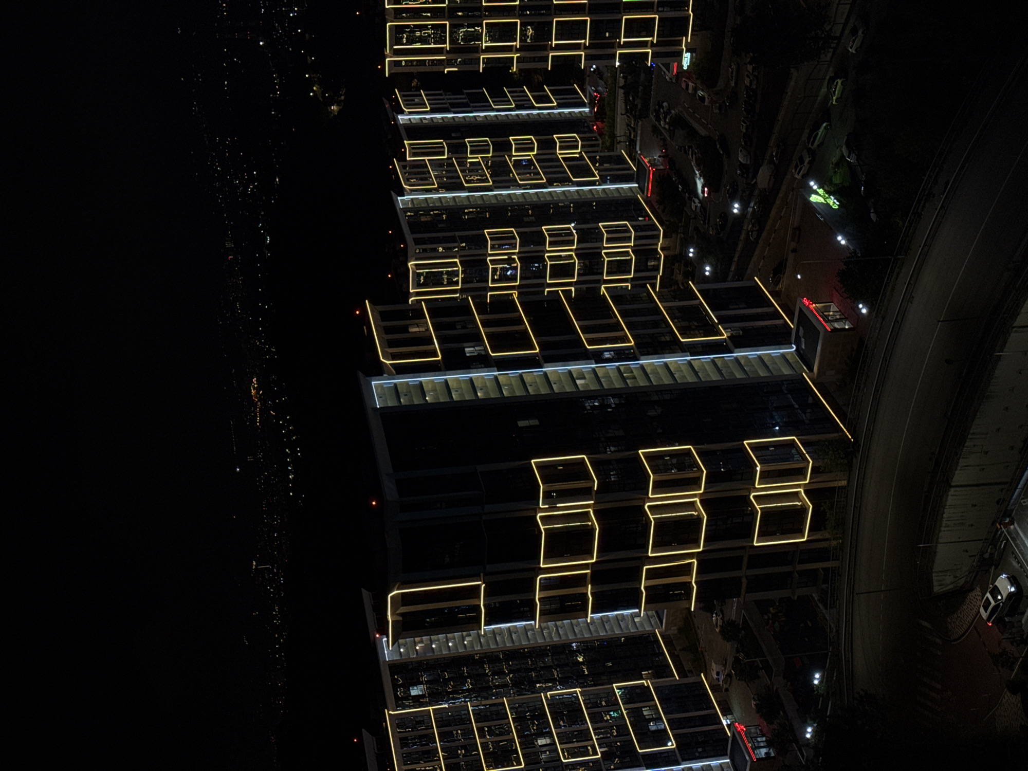 This is an aerial, night-time photograph of a modern residential complex in Sochi, Russia. The complex consists of several tall, multi-story buildings characterized by their dark facades and prominent yellow LED lighting that outlines the windows and architectural features. The lighting creates a striking geometric pattern against the dark buildings.
In the foreground, a curved road is visible, with a white SUV parked on it. Further down the road, there are several other cars parked in a lot. The immediate area around the buildings includes trees and some smaller structures, one with a red illuminated sign. In the background, a sprawling cityscape with numerous distant lights stretches across the horizon, indicating a large urban area. The sky is dark and appears clear. The overall impression is one of a contemporary urban development at night, with a focus on the illuminated architecture.