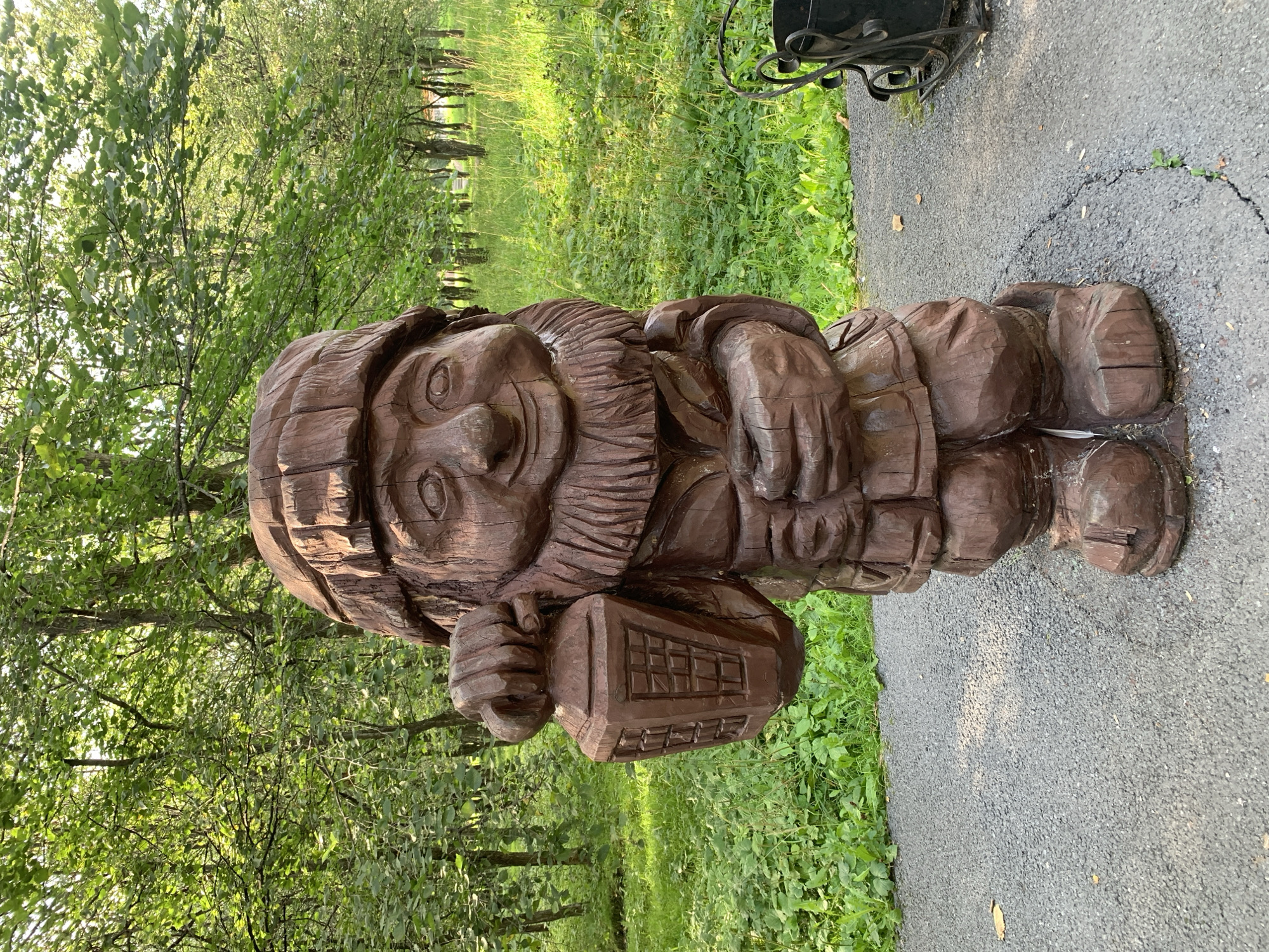 A wooden sculpture of a garden gnome stands in a park. The gnome is carved with a smiling face, a long beard, and a hat. It is holding a lantern in its left hand. The gnome is standing on a gravel path, with green grass and trees in the background. The image is taken from a medium distance, with the gnome filling most of the frame. The lighting is natural, with sunlight filtering through the trees. The sculpture is located in the city of Prokopyevsk, Russia, specifically in the Vinogradova Park, also known as the Park of Culture and Recreation.