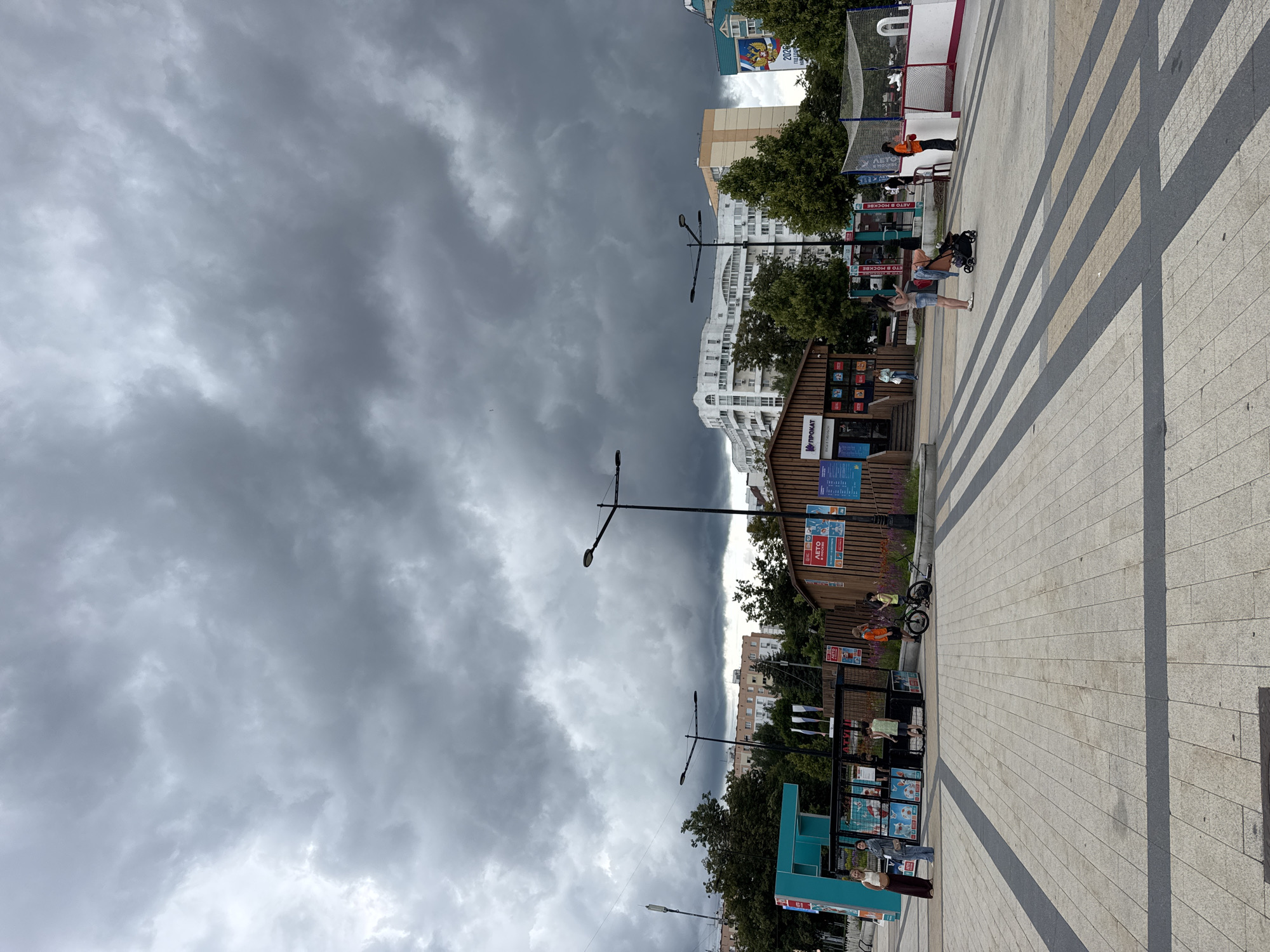 In Zelenograd, Russia, on a cloudy day with dark, dramatic storm clouds filling the sky, an open public square is depicted. The square's ground is paved with light-colored square tiles and features dark grey diagonal stripes. In the mid-ground, a wooden kiosk with signs is situated, with several people around it. To the left of the kiosk, a turquoise structure with glass panels displays posters and is attended by a woman in a light-colored dress and a woman in a grey outfit. Behind the kiosk, a chain-link fence encloses an area with netting, possibly a sports court, where people are visible. Trees line the background, with multi-story residential buildings visible beyond them, some featuring white facades and others with beige and green accents. Streetlights are present in the scene. A young child on a bicycle and a scooter are in the foreground near the kiosk. Further to the right, a woman stands near a stroller. Another woman in an orange shirt stands by the sports court area.