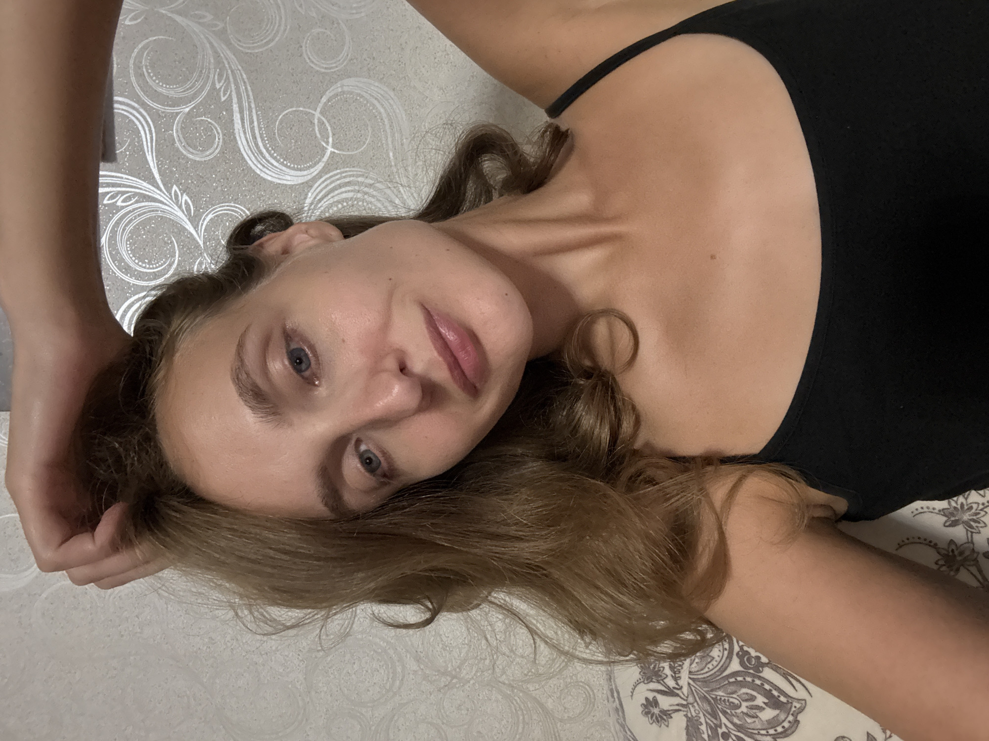 A young woman with light brown hair, blue eyes, and fair skin is captured in a selfie. She is wearing a black tank top and has her right arm raised, with her hand resting on her head. The background features a textured, light-colored wallpaper with a white floral pattern. A portion of a patterned pillow is visible in the lower left corner. The lighting suggests an indoor setting. The specific location in Kuyeda, Russia is not discernible from the image itself.