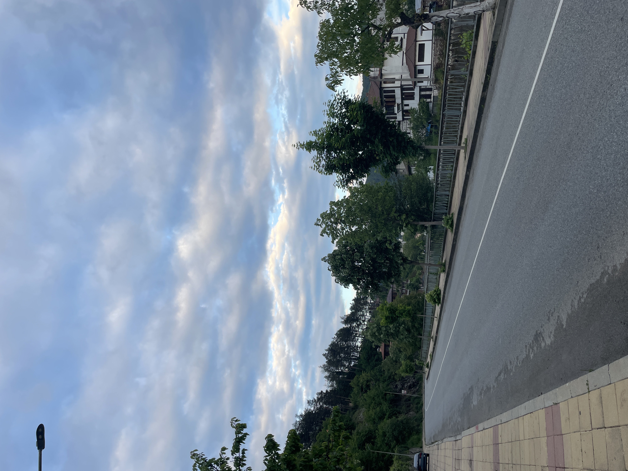 The image shows a street in Zlatograd, Bulgaria, with the sky taking up a large portion of the frame. The sky is filled with clouds with some blue showing through. On the right side of the image, there is a road with a white line dividing it. To the right of the road is a sidewalk made of yellow and pink stones. On the left side of the road is a metal fence, trees, and some buildings. The vegetation appears lush and green. A lamppost is visible in the lower left corner.