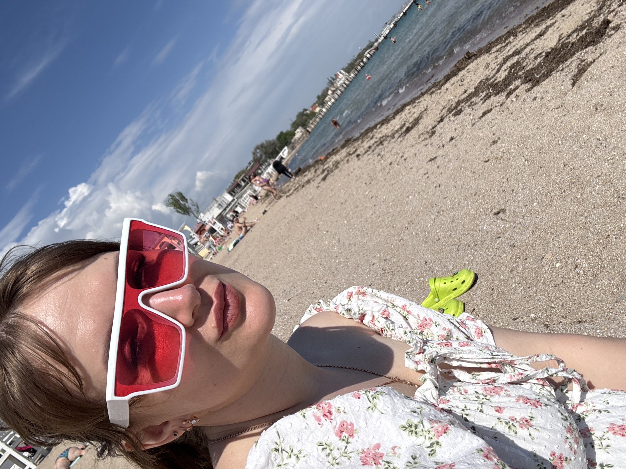 A woman with brown hair is lying on a sandy beach, wearing large white-framed sunglasses with red lenses and a white floral-print dress. She is positioned in the foreground of the image. A pair of lime green Crocs are next to her. The beach extends into the background, with other people visible sunbathing, walking, and swimming in the water. In the distance, there are buildings and structures along the coastline. The sky is blue with some clouds. The image appears to be taken at a beach in Yevpatoriya, Ukraine.