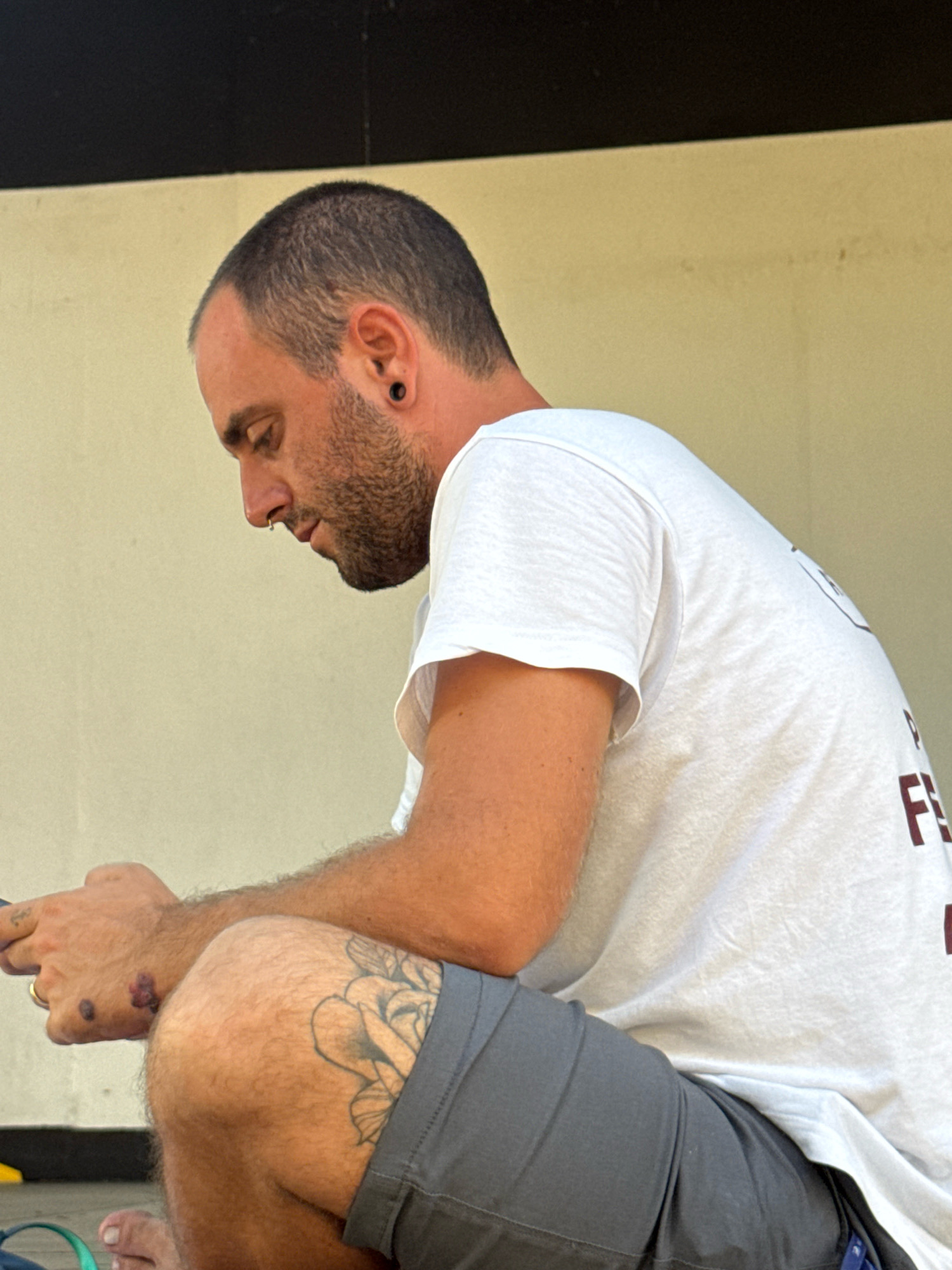 A man with a shaved head and beard is sitting outdoors, looking down at his phone. He is wearing a white t-shirt and grey shorts. He has a tattoo of a rose on his left thigh and several markings on his right hand. The background appears to be a wall. The image does not provide enough information to determine the specific location in Isola delle Femmine, Italy.