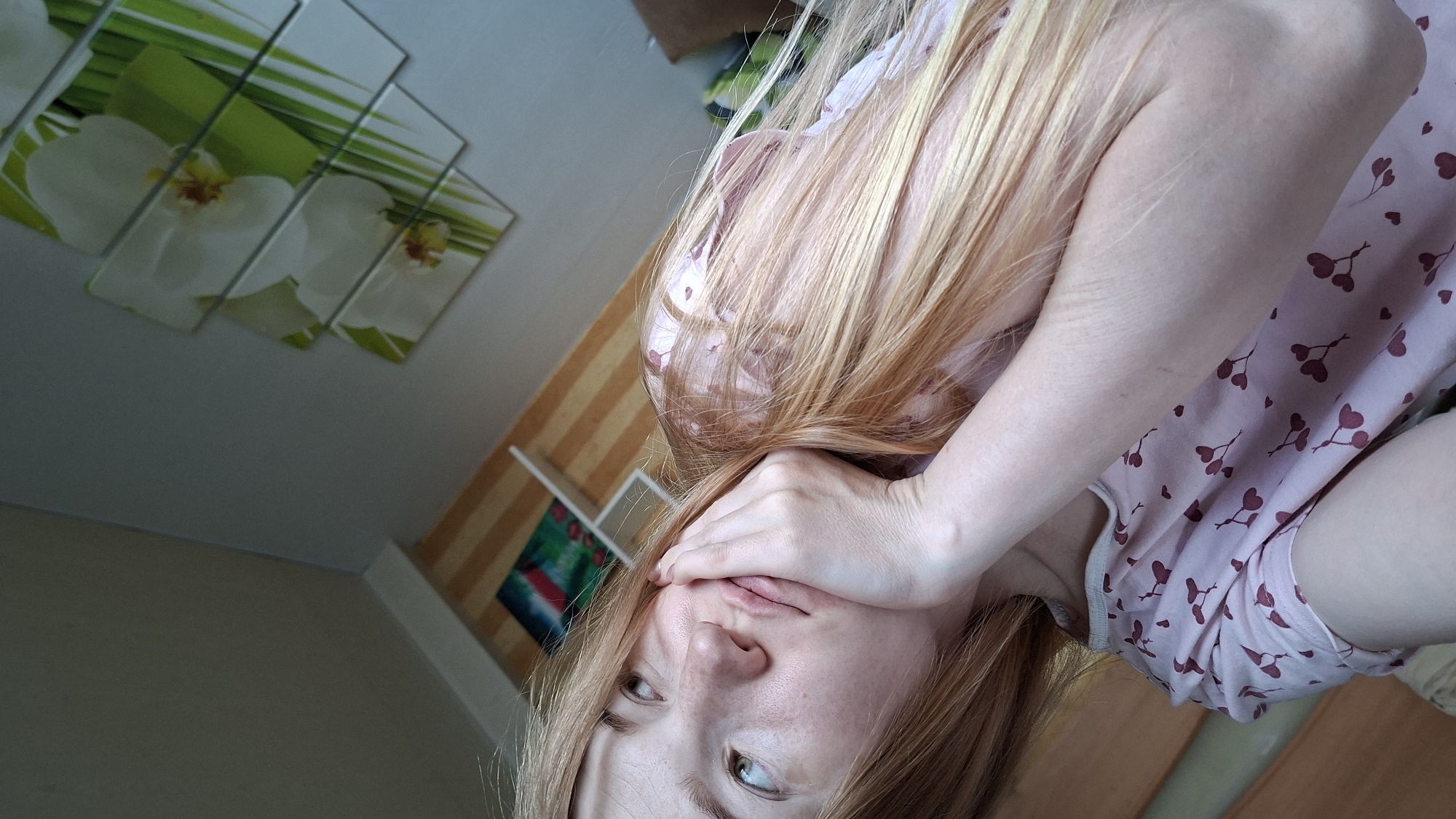 A woman with long, light blonde hair is reclining in an interior setting. She is wearing a light pink top with a pattern of small, dark red hearts. She is partially obscuring her face with her left hand. The environment includes a wall adorned with a multi-paneled artwork featuring white orchids and green foliage. Adjacent to the artwork, there is a shelf with various items, including what appears to be an abstract painting with red and blue hues. The location is Большой Букор, Russia, as indicated by the prompt.