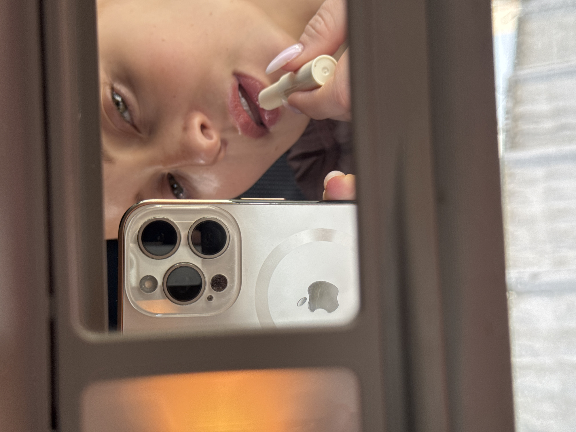 The image is a selfie taken in a mirror using an iPhone. The phone is a gold/white color and features a prominent Apple logo. The camera lenses are visible. The reflection shows a person applying lip balm to their lips. The person has light skin, visible eye, and manicured nails. The background is blurry and indistinct, potentially showing a window with light coming through. The scene location information is not contained within the image.