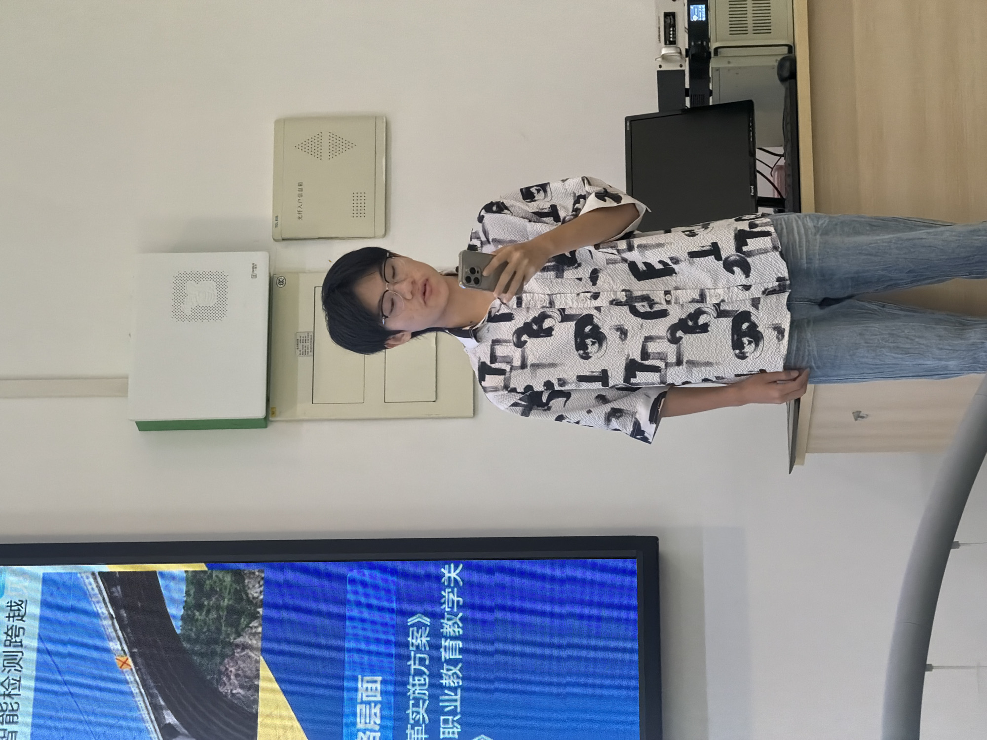 A young East Asian person wearing glasses, a patterned button-down shirt, and light blue jeans stands in front of a white wall and a large digital screen. The person is holding a smartphone in their right hand and appears to be looking at it. The digital screen on the left displays an image of a bridge and Chinese text. Mounted on the wall are several utility boxes. To the right of the person, on a desk, are computer equipment, including a monitor and what appears to be audio or communication devices. The lighting suggests an indoor setting, possibly a classroom or presentation room. The overall scene appears to be at a vocational or technical school in Jiangjin, China, as indicated by the text on the screen and the context of the setting.