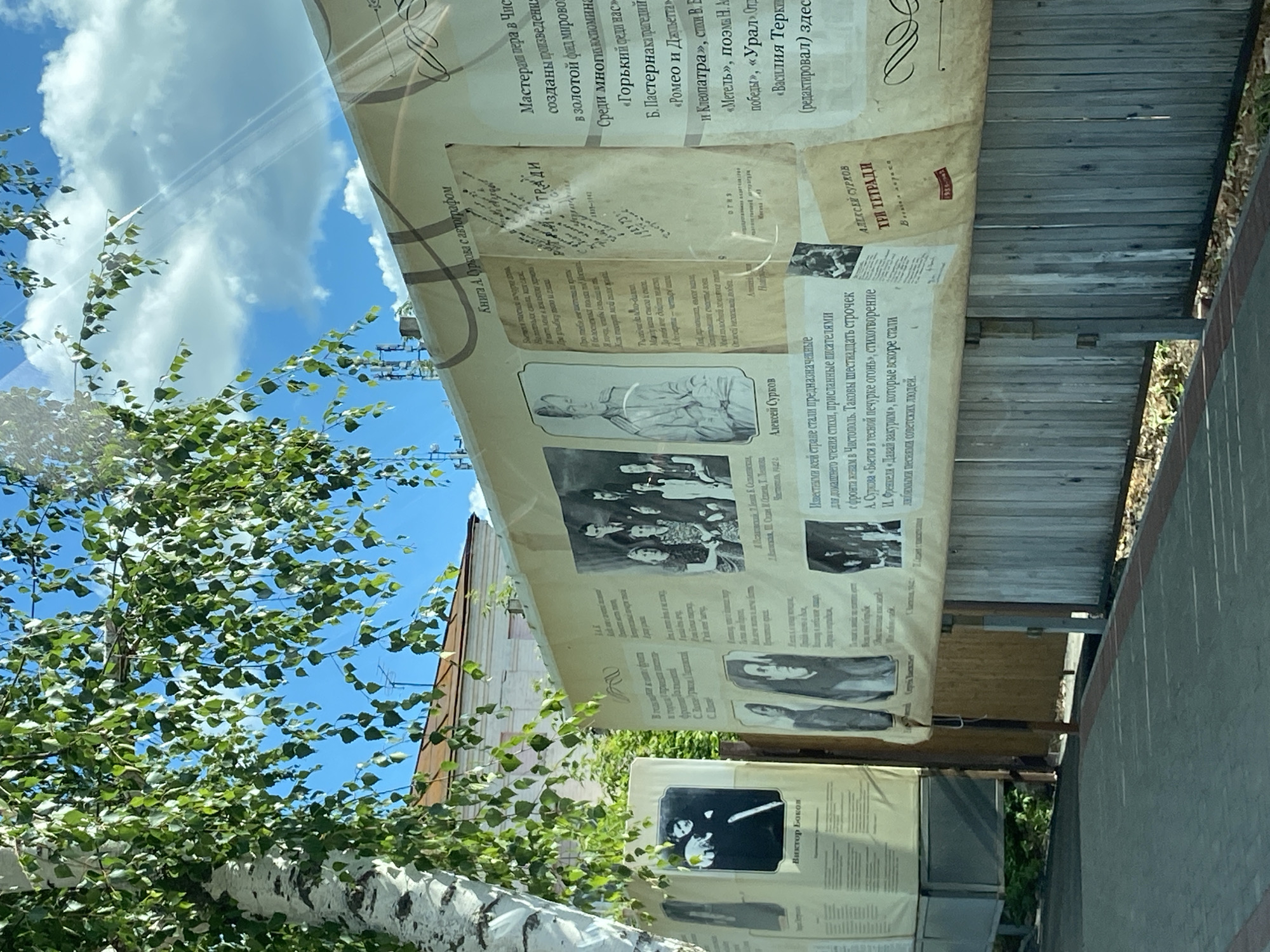 An outdoor scene captured at a slightly tilted angle, showing a bright, sunny day with a clear blue sky dotted with fluffy white clouds. The primary subjects are large, parchment-like banners, possibly made of canvas or fabric, hanging vertically. These banners serve as an informational display, featuring text and black-and-white photographs of individuals, suggesting a historical or literary exhibition.
In the foreground, the vibrant green leaves of a birch tree are visible, with its distinctive white and black mottled trunk prominent on the left. The tree's branches and foliage partially obscure the sky and parts of the banners, indicating they are positioned outdoors, perhaps in a park or along a street.
The banners display biographical information and excerpts from literary works, primarily in Russian. Notable text includes mentions of "Мастерами пера в Чистополе" (Masters of the pen in Chistopol), indicating the location context of Chistopol, Russia. Names like "Алексей Сурков" (Aleksey Surkov) and "Виктор Боков" (Viktor Bokov) are clearly visible, accompanied by their portraits. One section details poems sent by writers from the front to their wives in Chistopol, specifically mentioning A. Surkov's "Бьется в тесной печурке огонь" (Fire burns in a cramped stove) and I. Frenkel's "Давай закурим" (Let's light up a cigarette), which became popular Soviet songs. Other literary references include "Горький фели нас," "Б. Пастернака трагедий," "Ромео и Дальетта," "Метель," and "Василия Теркин."
Behind the banners on the right, a weathered wooden fence suggests the boundary of a property or garden. The overall impression is that of an open-air museum or a temporary cultural display, designed to inform passersby about local literary history or significant figures from the region. The bright sunlight and clear sky contribute to a pleasant, inviting atmosphere.