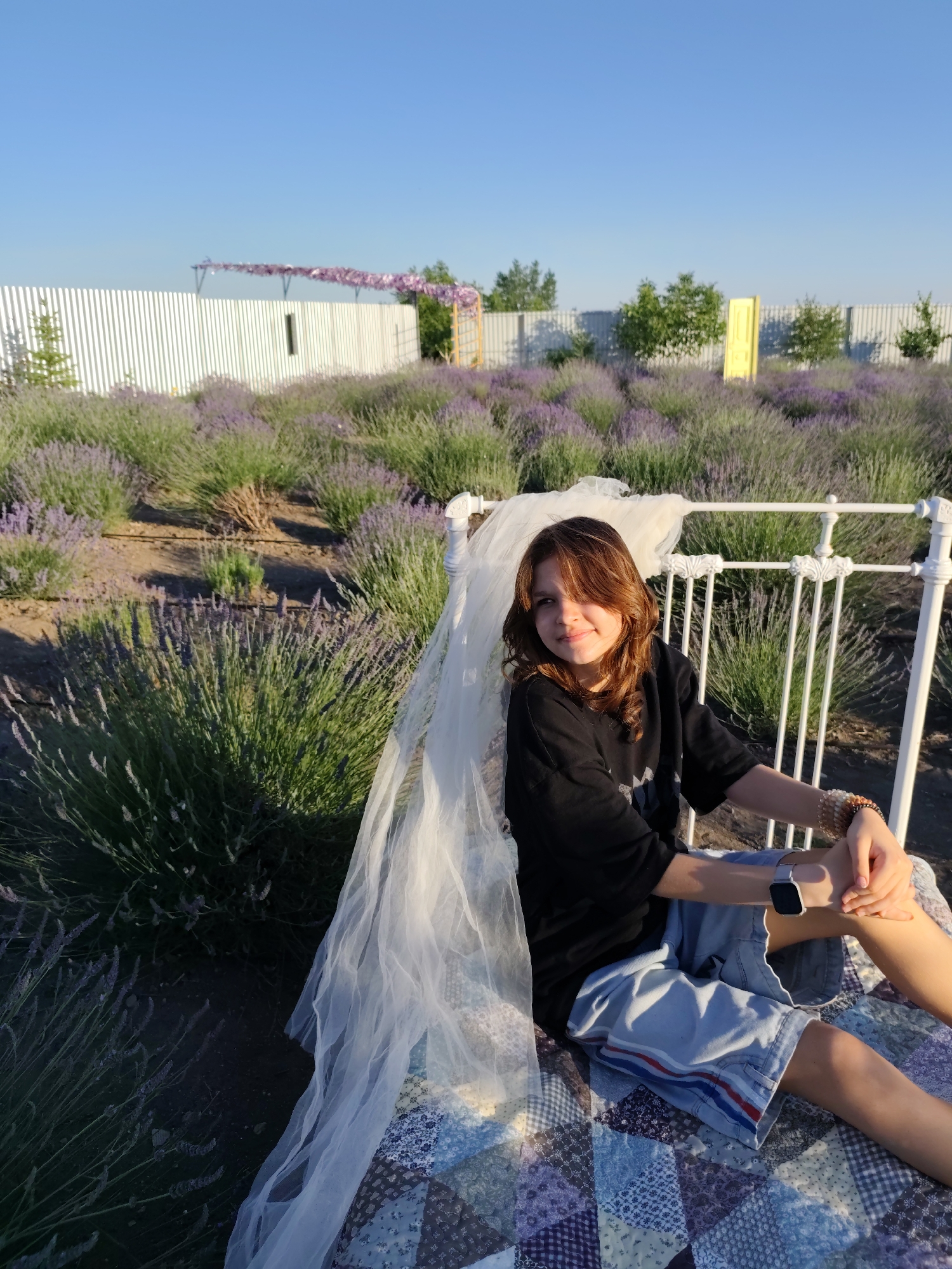 A young person with shoulder-length brown hair and light brown eyes is sitting on a white ornate bed frame in a lavender field. They are wearing a black t-shirt and light blue denim shorts with red and blue stripes on the side. A light white veil is draped over the bed frame behind and to the left of the person. The bed is covered with a colorful patchwork quilt. The person is looking at the camera with a slight smile, and their right arm is resting on their right leg, while their left arm is bent with their hand on their knee. A watch with a grey band is visible on their left wrist.
The scene is a lavender field under a clear blue sky. In the background, there is a white picket fence, some trees, and a decorative yellow door. The sun appears to be low in the sky, casting a warm glow on the scene, suggesting it is late afternoon or early evening. The overall mood is peaceful and idyllic.
Nechaiane, Ukraine is the stated location context.