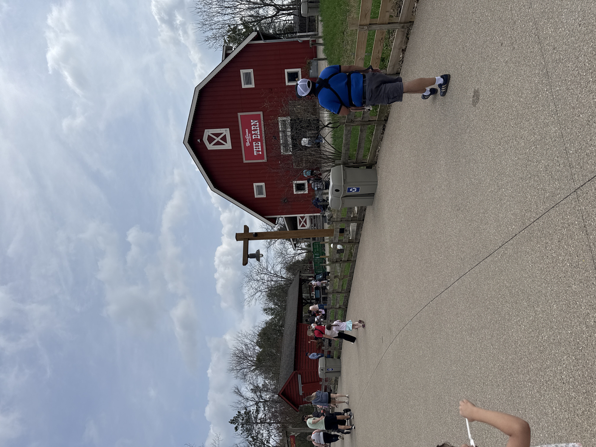 A public outdoor space near "The Barn" in Powell, Ohio, United States, shows a red barn building with white trim under a cloudy sky. A person in a blue shirt and white cap is seen walking along a paved path, next to a wooden fence. People are scattered throughout the area, some near another red building further down the path. Trash and recycling bins are present.