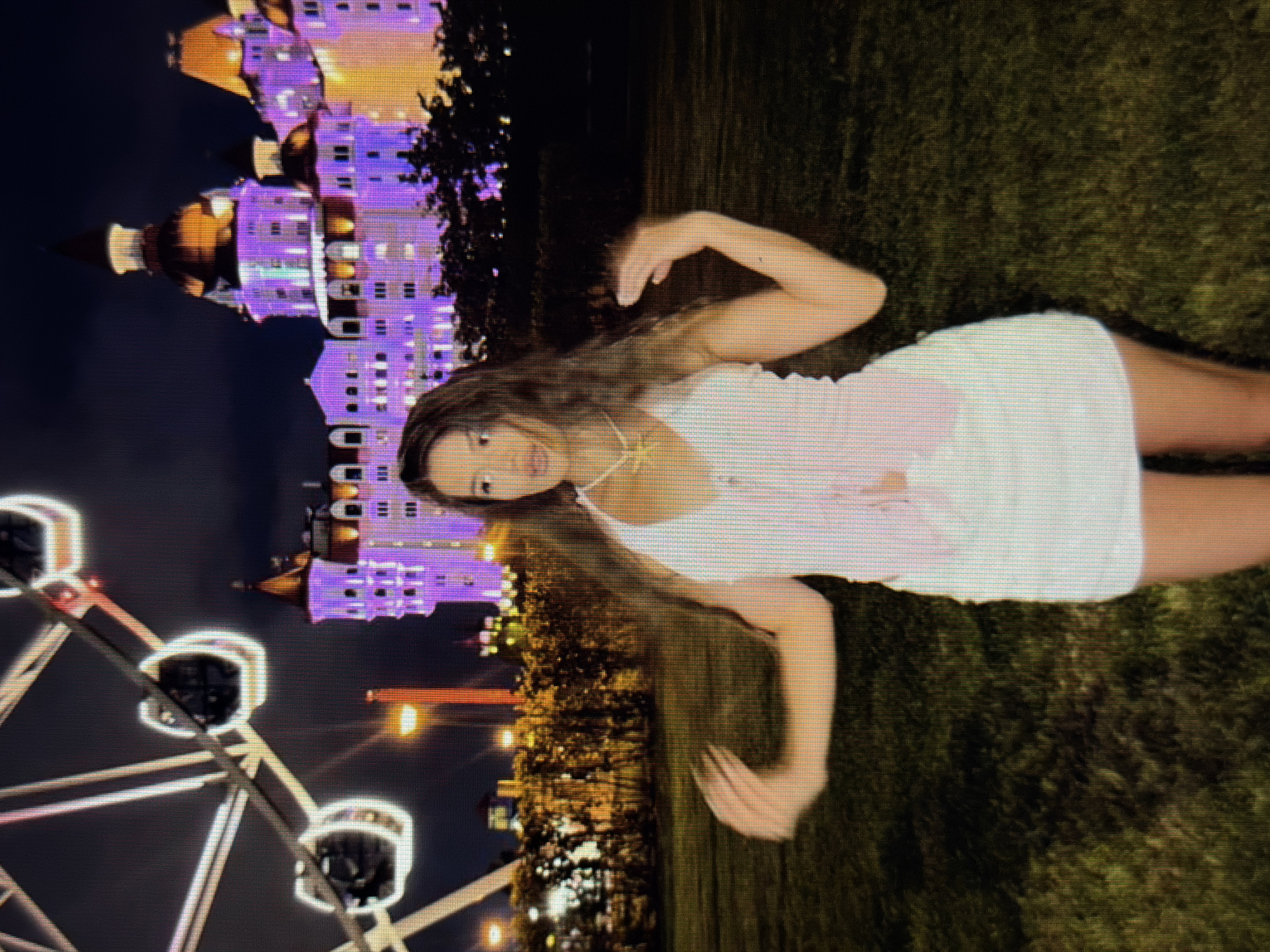 A young woman with long, dark, wavy hair stands in a grassy area at night. She is wearing a white halter top with pink trim and a white skirt. Her arms are outstretched, and her hands are open. Behind her, a large Ferris wheel with illuminated cabins is visible on the left side of the frame. To the right of the Ferris wheel is a large, ornate building with purple and orange lights illuminating its facade and domes. The building is likely the Ice Palace "Bolshoi" or "Maly" in Sirius, Russia. The sky is dark, indicating it is nighttime. The overall scene suggests an amusement park or entertainment district setting.
