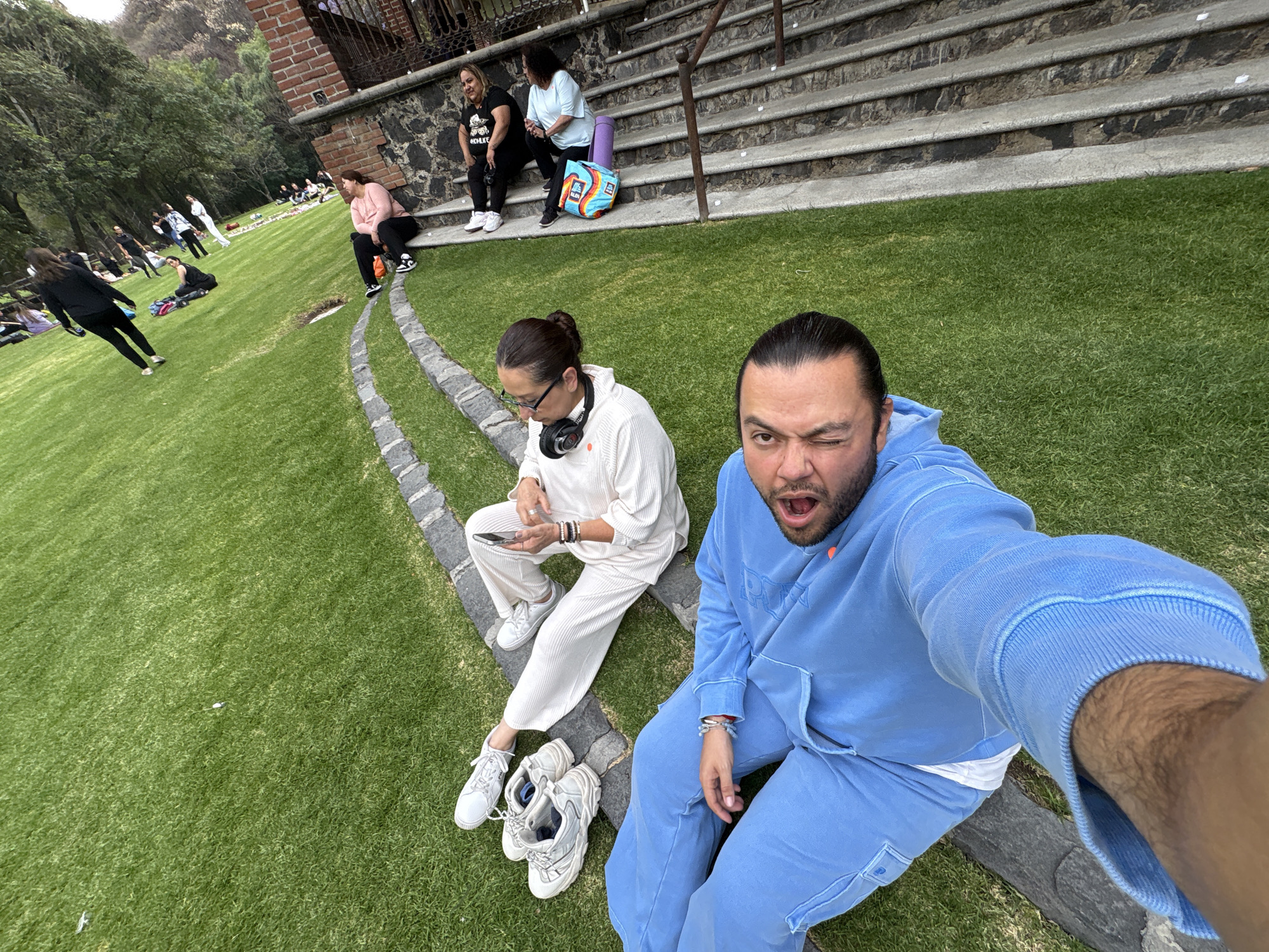 The image shows two individuals seated on a stone border in a grassy outdoor area. A man in a blue tracksuit is taking a selfie, with one eye closed and mouth open. Next to him, a woman wearing a white outfit and headphones is looking at a phone. Her sneakers are next to her. Behind them are people sitting on stone steps of a building. In the background, more people are visible on the lawn. The scene is in Santa Ana Acozautla, Mexico.