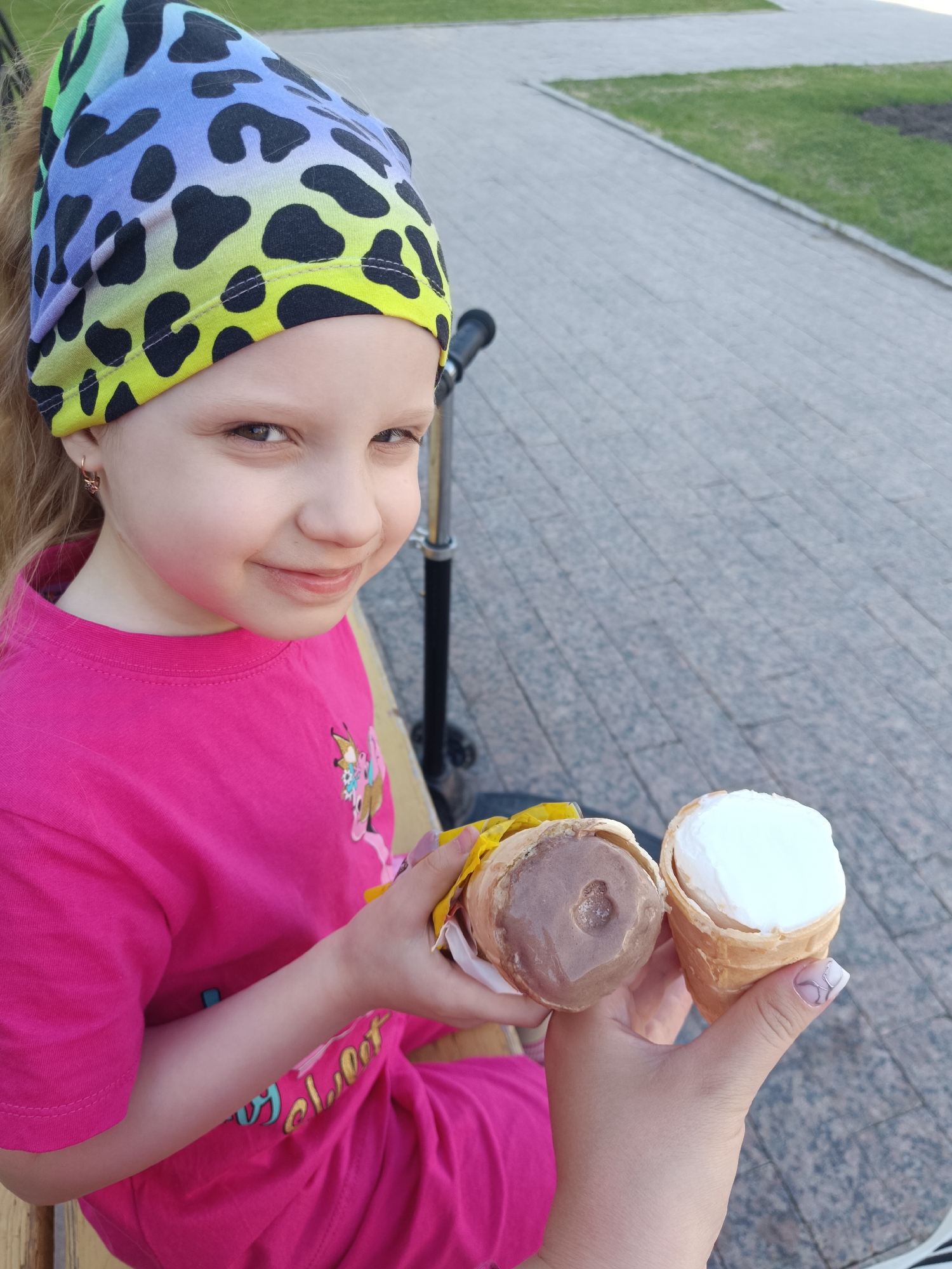 A young girl with a colorful bandana sits outside, holding two ice cream cones. The ice cream flavors are vanilla and chocolate. In the background, a scooter is visible, along with a paved walkway and patches of green grass. The setting is Tobolsk, Russia.