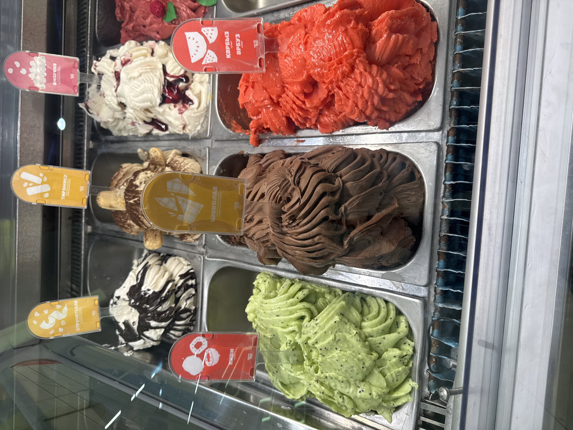 A close-up, eye-level shot displays several metal trays filled with vibrant ice cream flavors, presented in a refrigerated display case, likely in a shop. The scene is brightly lit, suggesting an indoor setting during daytime.
The trays contain an assortment of ice cream textures and colors: a deep red, a creamy white with swirls of dark red jam, a rich brown chocolate, a swirl of white and black, and a light green ice cream speckled with black seeds. Each tray has a small, colorful label inserted into the ice cream.
The visible text on the labels includes:
- A red label with white stylized watermelon slices and the text "КАРБЫЗ" and "АРБУЗ" in Cyrillic.
- A red label with white stylized fruit shapes and the text "ЯБЛОЧНАЯ" in Cyrillic.
- A yellow label with white stylized waves and the text "ТИРАМИСУ" in Cyrillic.
- A yellow label with white stylized swirls and the text "СТРЯПЧЕЛЯ" in Cyrillic.
- A red label with white stylized fruit shapes and the text "КИВИ" in Cyrillic.
The overall impression is one of a delicious, inviting selection of frozen treats, with the focus on the textures and colors of the ice cream. The setting appears to be a commercial establishment, possibly a gelato shop or ice cream parlor in Almaty, Kazakhstan, given the Cyrillic text. There are no people visible in the frame.