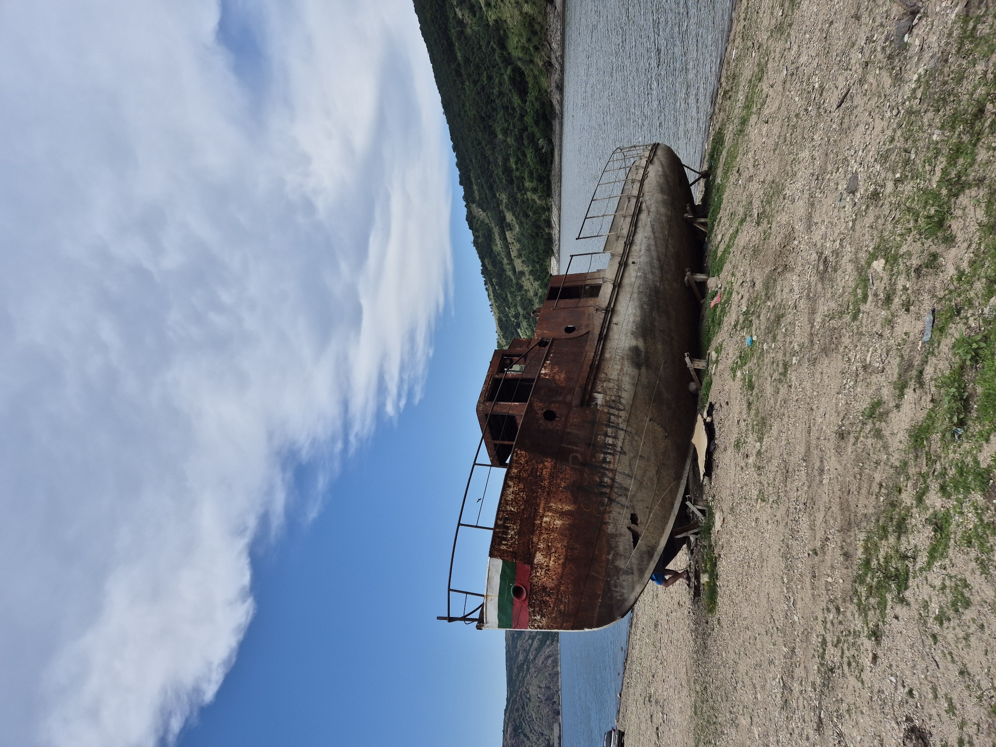 This forensic description details a large, derelict, rust-covered metal boat beached on a dry, sandy shore beside a body of water in Shiroko pole, Bulgaria. The vessel, oriented with its bow facing left, displays extensive orange-brown corrosion across its entire hull and superstructure. The bow is painted with faded horizontal stripes of white, green, and red, indicative of the Bulgarian flag. Visible graffiti or markings are present on the rusted hull. The boat is supported by several wooden chocks and planks, stabilizing its tilted position on the shore. Partially visible beneath the bow, the lower legs and feet of an individual, clad in blue shorts and dark sandals, suggest a person is positioned under the hull, possibly inspecting or working on it.
The immediate foreground consists of dry, light-brown soil mixed with pebbles and sparse patches of green grass, with some scattered debris. To the right of the boat, the calm surface of a large lake or reservoir extends towards forested, undulating hills that form the distant background. The hills are covered with dense green foliage and lighter patches of vegetation. In the far left background, a dark-colored car is parked near the water's edge. The sky above is largely clear and blue, with prominent white cumulus clouds distributed across the upper half of the frame.
