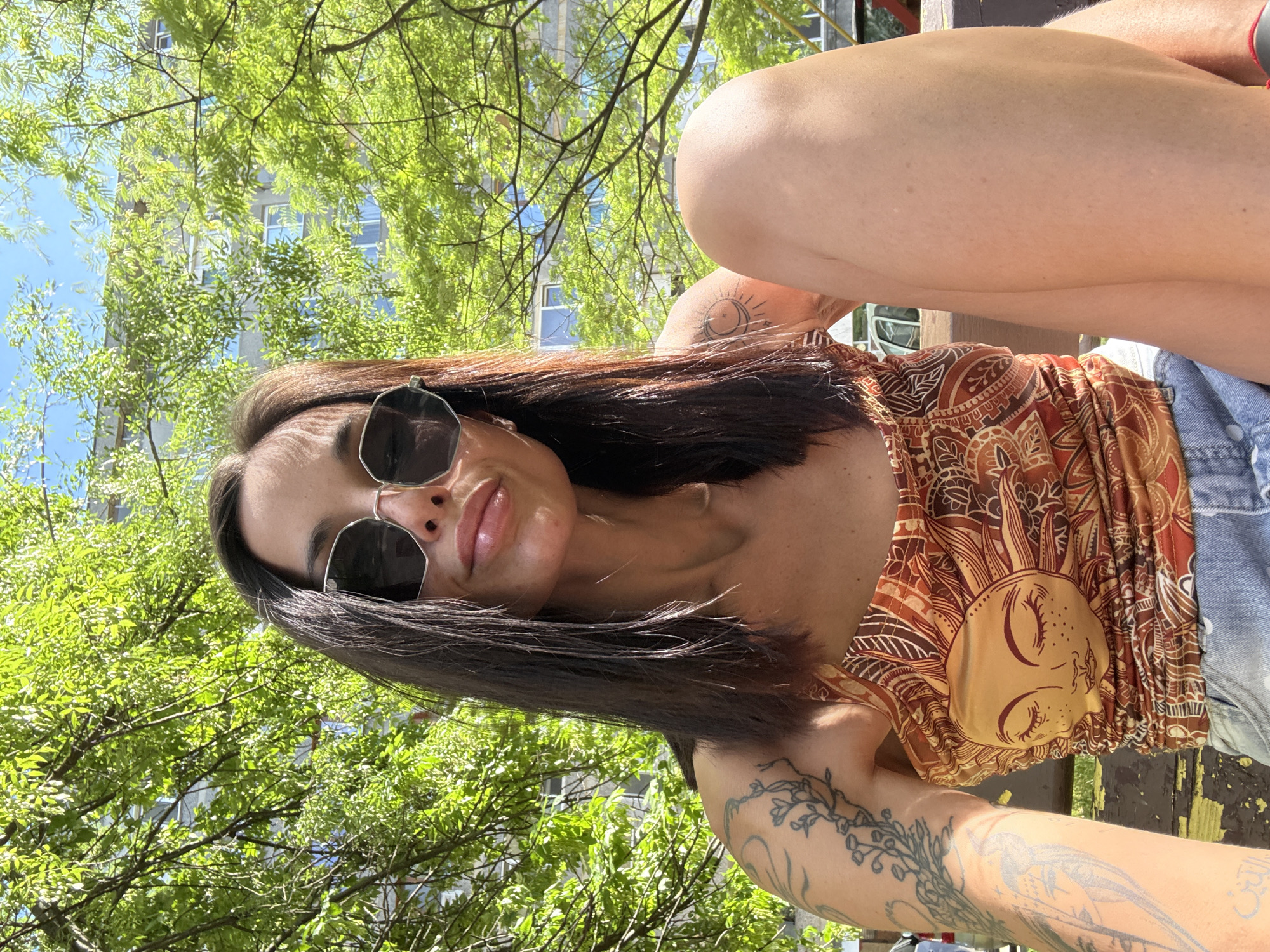 A woman with dark hair and sunglasses is posing for a photo outdoors. She is wearing a patterned top with a sun design and denim shorts. She has tattoos on her arms and chest. The background includes green trees, a building, and a blue sky. The location is possibly in Pleven, Bulgaria.