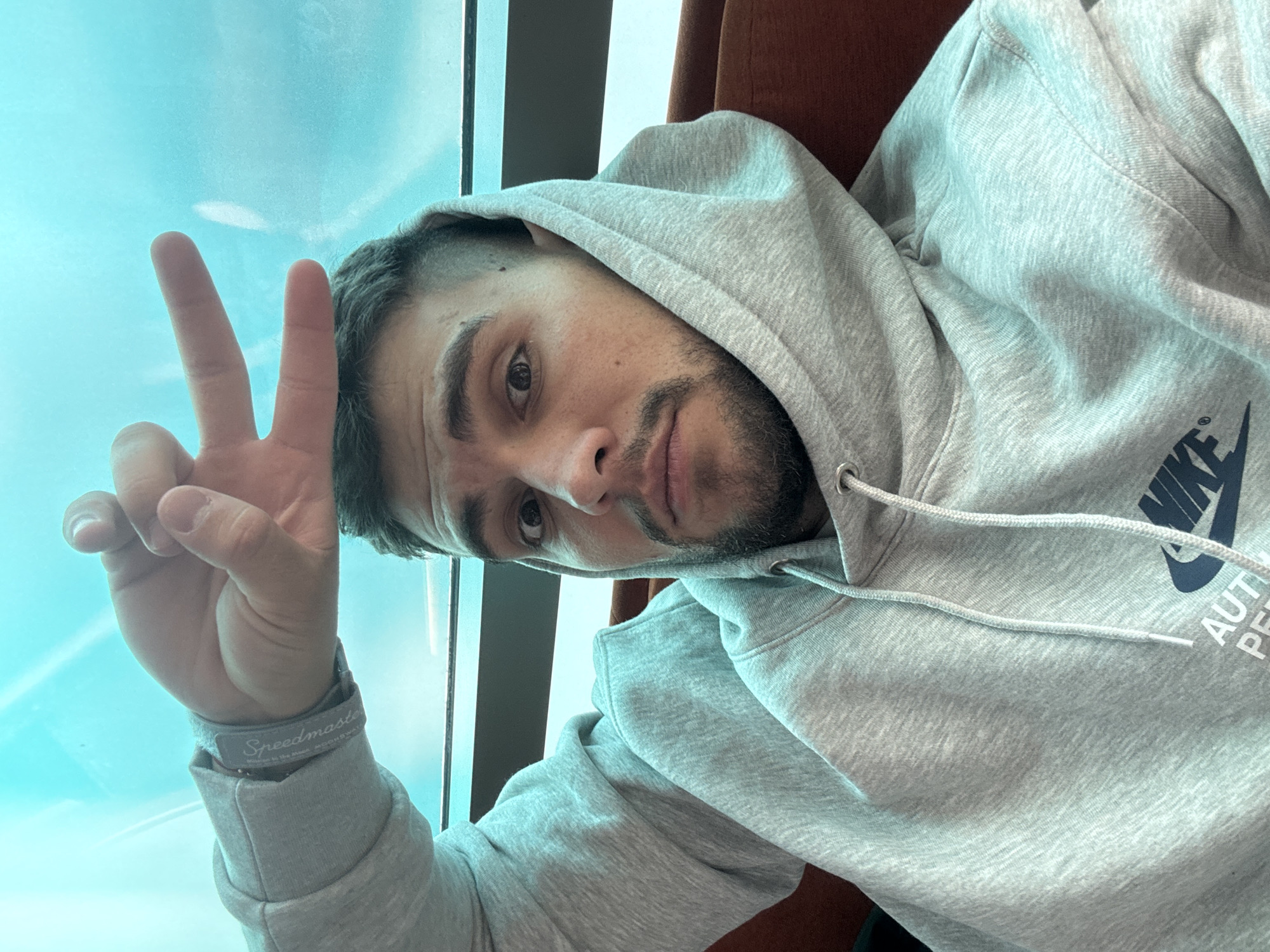 A man is seated next to a window, possibly indoors, based on the window frame and the light. He is wearing a gray hooded sweatshirt with a "Nike" logo visible on the right side. The man is making a "peace" or "victory" sign with his left hand. He has short dark hair, a trimmed beard, and fair skin. A "Speedmaster" wristband is visible on his left wrist.
Based on the light conditions, the location is likely indoors during daytime. No landmarks that would allow identifying the location in Sofia, Bulgaria, are visible in the scene.