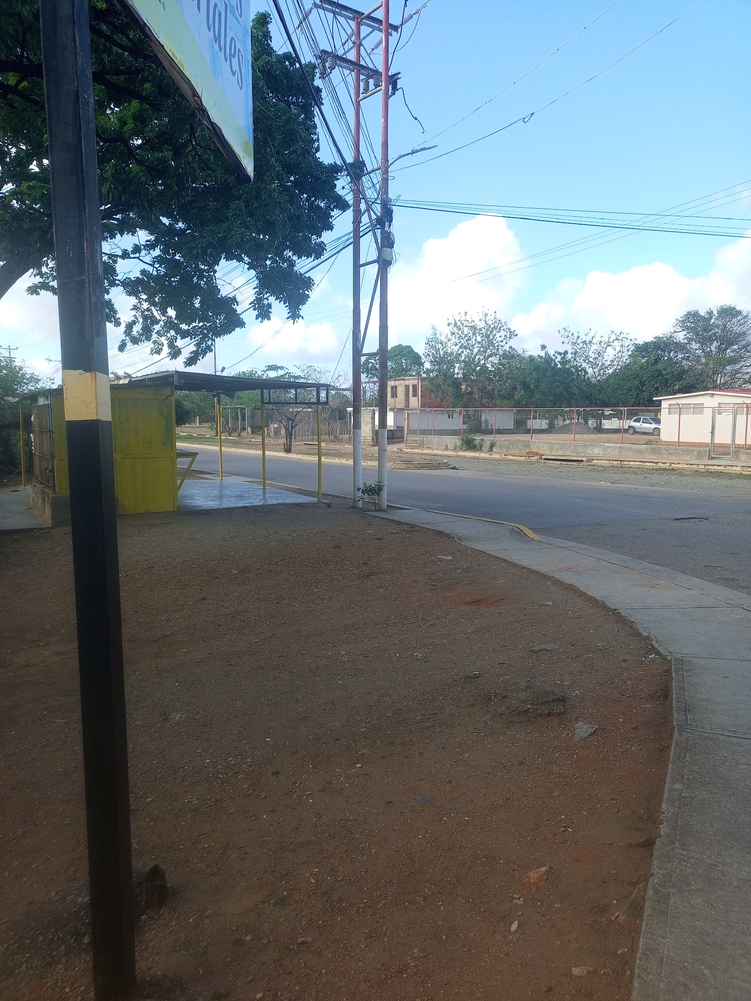 The image depicts a street scene in Altagracia, Venezuela, featuring a bus stop shelter. The shelter has a yellow metal structure with a corrugated metal roof, supported by yellow poles. The ground in front of the shelter is a mix of dirt and gravel. To the left of the shelter, a dark pole with yellow accents stands prominently. In the background, a paved road curves to the right, with a sidewalk and a dirt shoulder. Across the road, there is a fenced area with several buildings, including a white structure with a red roof. Trees and utility poles with numerous wires are visible throughout the scene. The sky is partly cloudy. There are no people or animals visible in the image.