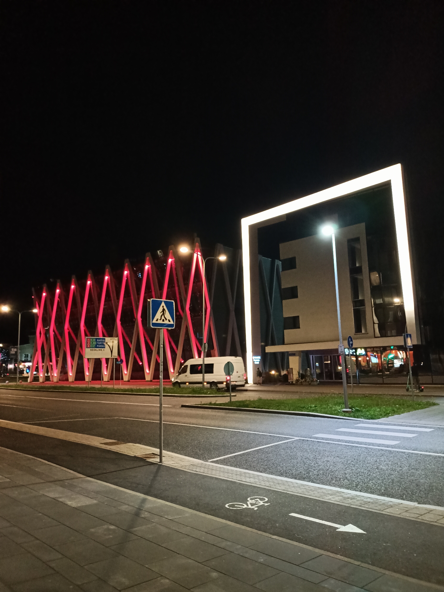 This is a nighttime, eye-level, medium shot of a street in Tallinn, Estonia. The scene is dominated by modern architecture and illuminated by artificial lights. On the left, a building with a distinctive diamond-patterned facade is lit with bright red lights. To the right, a larger, rectangular building is framed by a large, illuminated white border. The building itself is a lighter neutral color with dark windows and appears to be a multi-story structure with a commercial ground floor. In front of this building, a white van is parked on the street. A pedestrian crossing and a bicycle lane are clearly marked on the asphalt in the foreground, indicating a well-developed urban infrastructure. The sky is dark, with no celestial bodies visible, and streetlights are present, casting illumination on the road and sidewalk. A traffic sign indicating a pedestrian crossing is visible near the red-lit building.