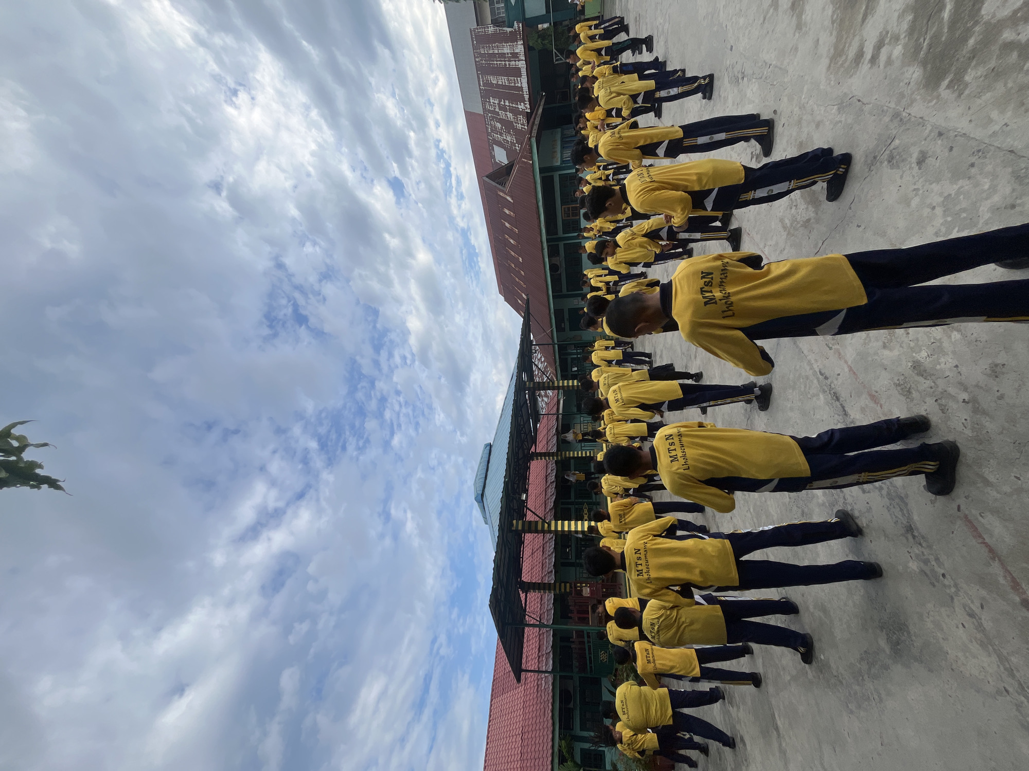 An outdoor, low-angle, full shot shows a group of male students in yellow shirts and navy pants with yellow stripes standing in a schoolyard. The students are facing away from the camera and appear to be engaged in some form of physical activity or assembly. The school building is visible in the background, with a red roof and green walls. The sky is cloudy with patches of blue. The location is identified as MTsN Lhokseumawe, a public junior high school in Lhokseumawe, Indonesia. The students' shirts have the text "MTsN Lhokseumawe" printed on the back.