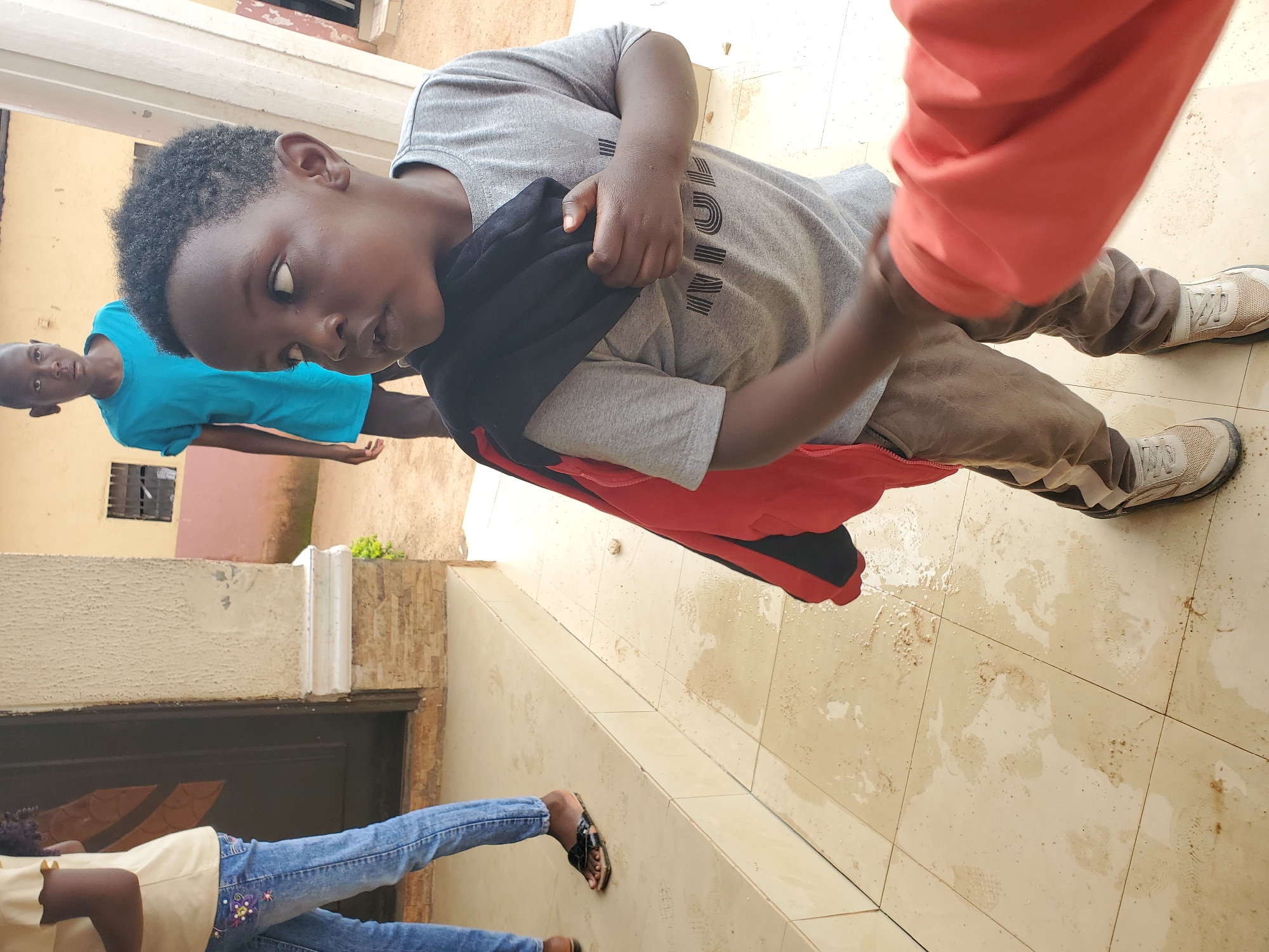A medium shot captures a young Black boy with dark curly hair, wearing a gray t-shirt, brown pants, and light-colored sneakers, standing outdoors on a tiled floor. He is holding a black and red jacket over his left shoulder and arm, with his right hand gripping the jacket. His gaze is directed to his right, with his mouth slightly open. Behind him and to his left, another young Black boy in a bright blue t-shirt and dark pants stands looking forward. To the far left, partially visible, is a person wearing a light yellow shirt and blue jeans. The setting appears to be the exterior of a building with a cream-colored textured wall and a dark doorway. The tiled floor shows signs of wetness and dirt. The overall scene suggests a casual outdoor environment, possibly residential, in the city of Jos, Nigeria.