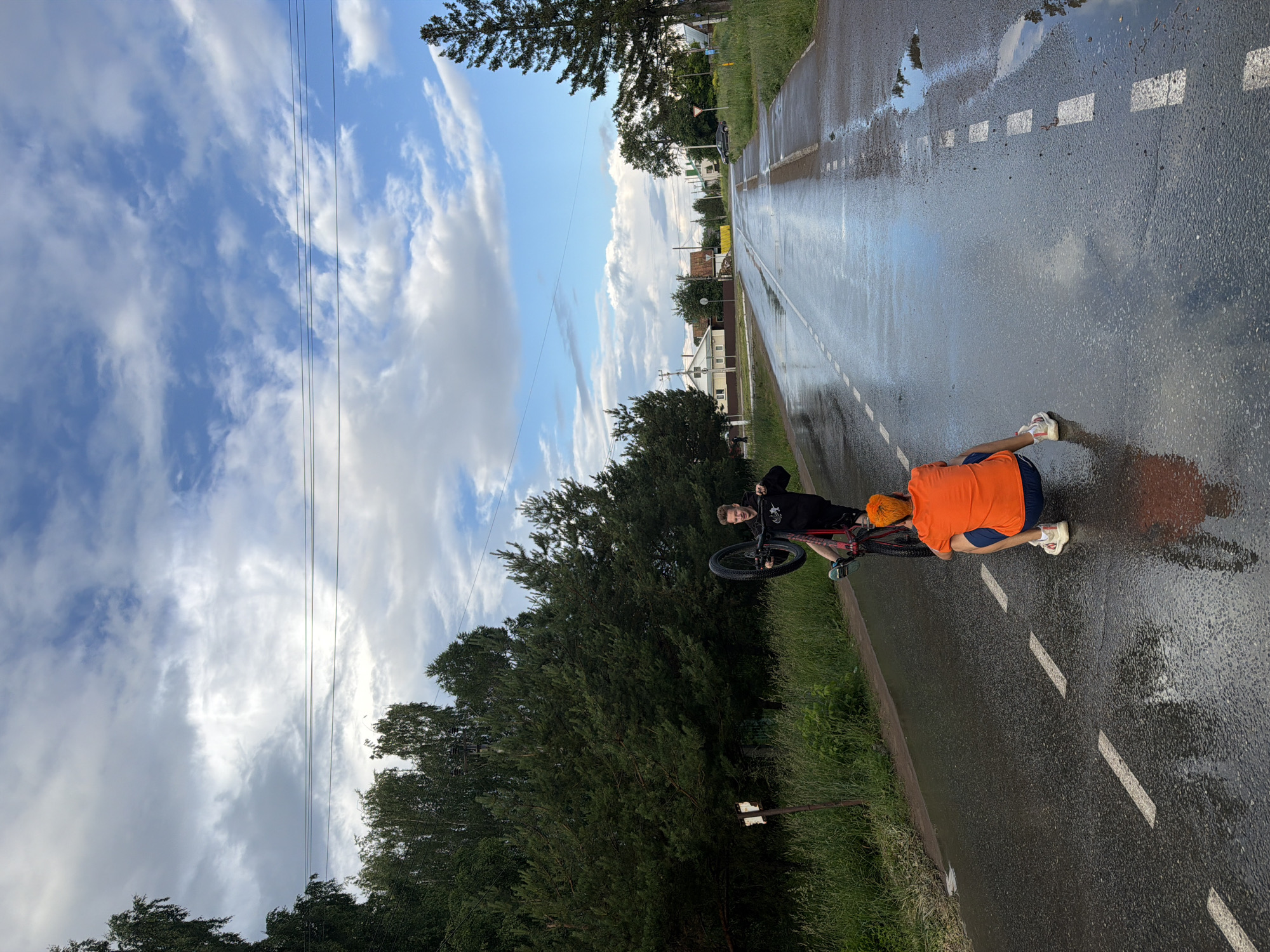 The image depicts two young males on a wet asphalt road, likely in Chaikovsky, Russia. One male is performing a wheelie on a red bicycle, with the front wheel raised. He is wearing a black hooded sweatshirt and dark shorts. The other male is crouched down on the road, facing away from the camera and towards the cyclist. He is wearing a bright orange t-shirt and dark blue shorts, with white athletic shoes. The road is wet and reflects the cloudy sky, suggesting it has recently rained. Trees and grass line the sides of the road, and in the background, houses and a few more trees are visible, indicating a rural or suburban setting. The sky is partly cloudy with patches of blue sky.