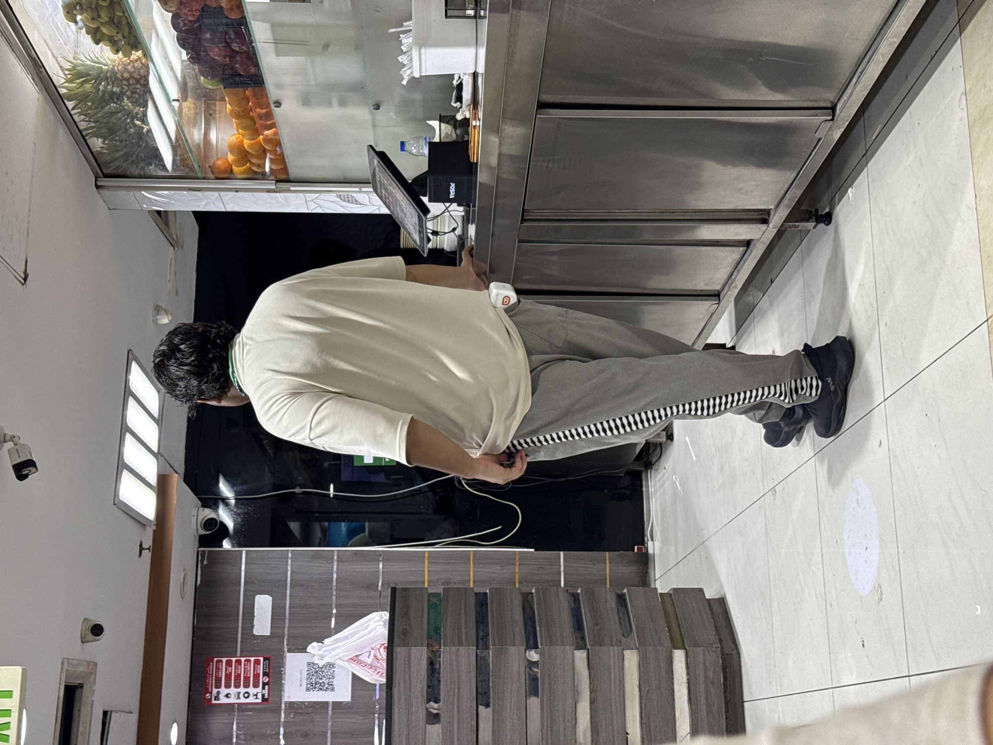 The image shows the interior of a juice or food establishment in Dubai, United Arab Emirates. The scene is viewed from behind a man standing at a stainless steel counter. The man is wearing a light-colored t-shirt and grey sweatpants with a black and white striped pattern down the side. He is facing towards the counter, which has a touchscreen display for a point-of-sale system, a bottle of water, and containers of utensils. Behind the counter, a glass display case showcases various fruits, including pineapples, oranges, and apples. To the left of the man, a counter or shelving unit is stacked with items wrapped in a grey, textured material. A white plastic bag and a QR code are visible on this unit. Several security cameras are mounted on the ceiling. The floor is tiled in a light color. The overall environment appears to be a retail space or service counter.