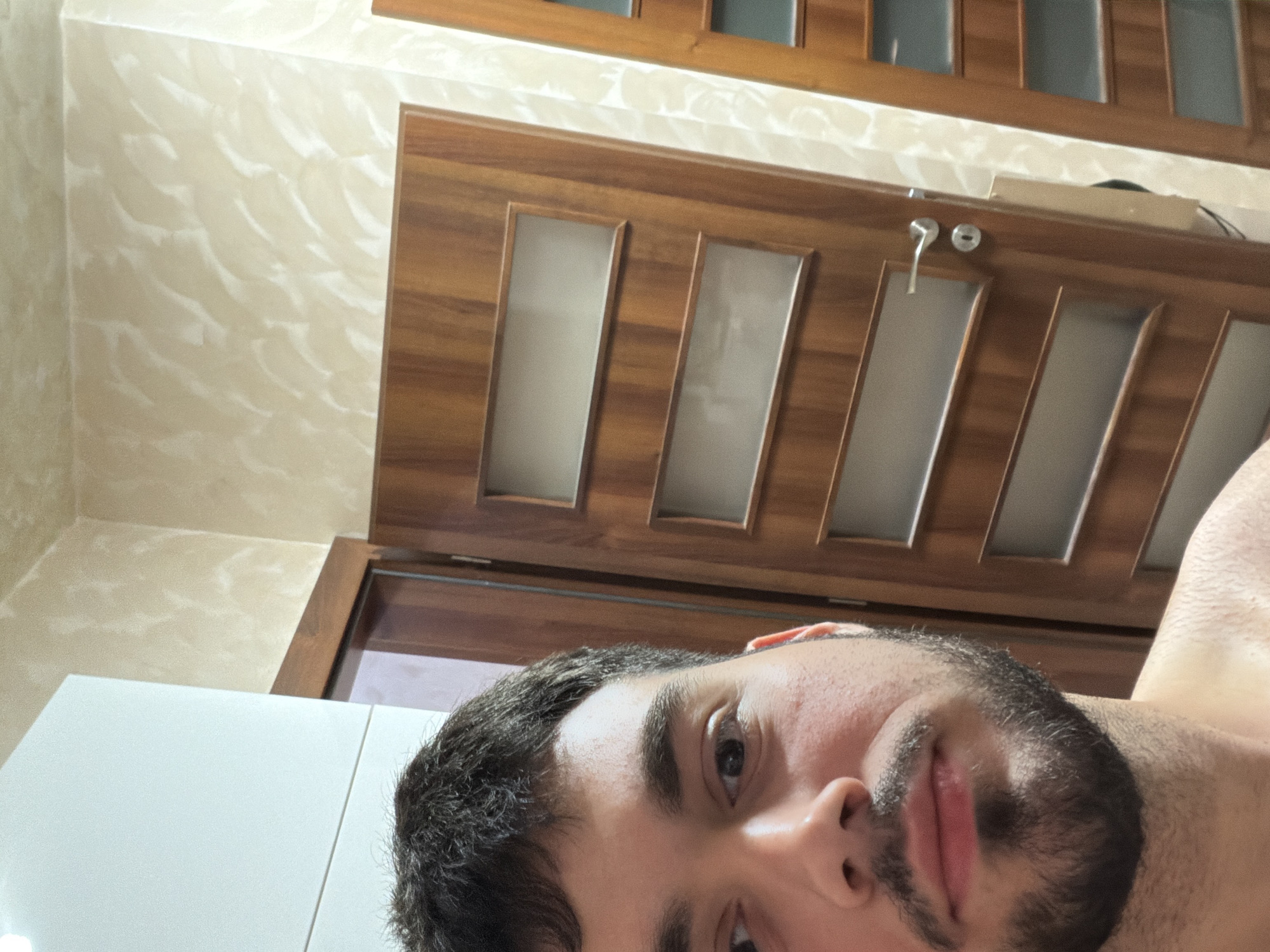 The image captures a selfie taken in what appears to be a residential interior. The foreground is dominated by the face and upper torso of a young man with short dark hair and a beard. He is looking directly at the camera. The background reveals an interior environment with two wooden doors. The door on the right is partially open, showing a glimpse of another door or wall beyond. The walls have a textured, light-colored finish. The overall scene suggests a personal space, possibly a hallway or entrance area within a home in the city of Sofia, Bulgaria.