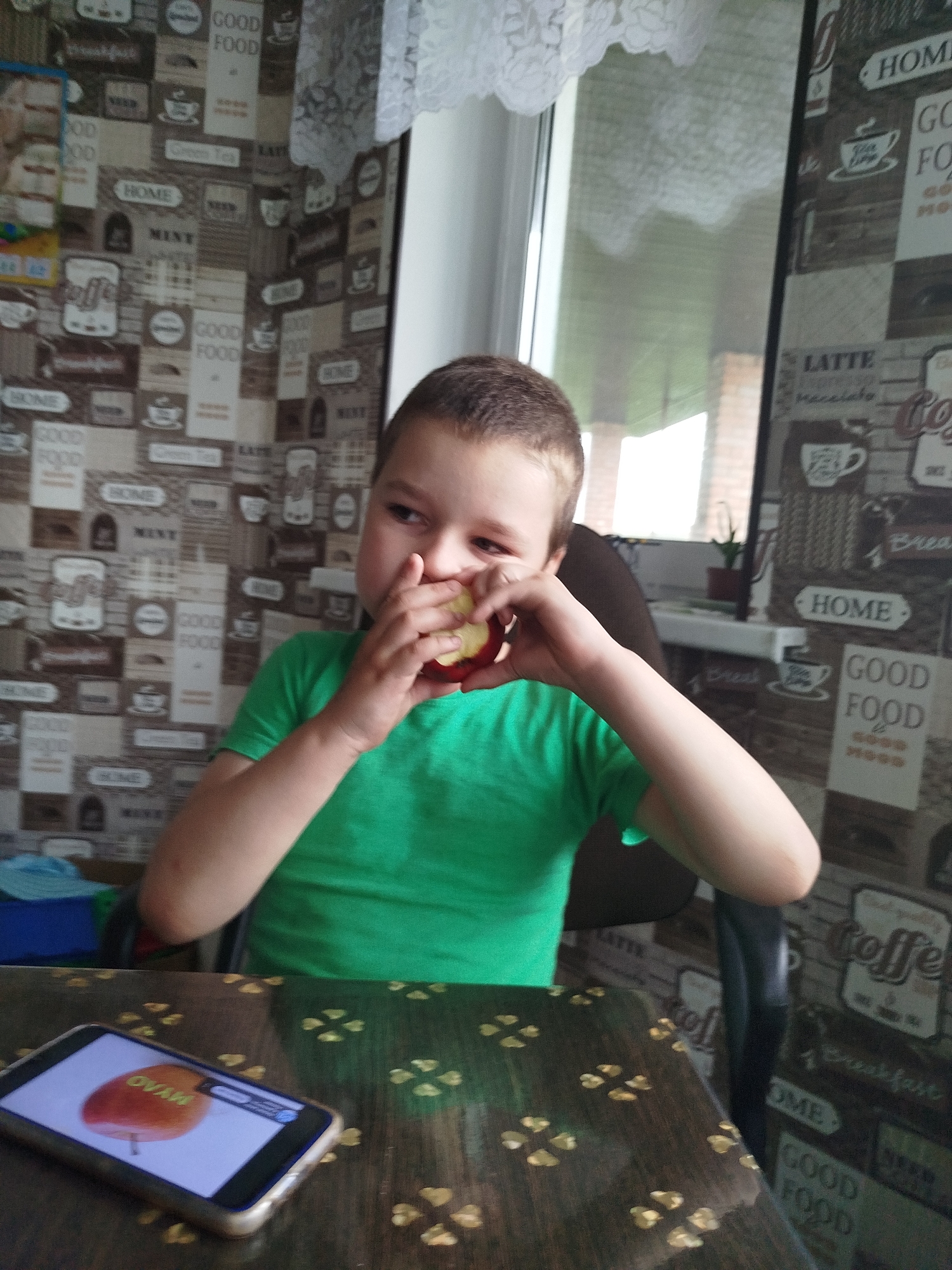 A young boy wearing a green t-shirt is sitting at a table, eating an apple. He is holding the apple with both hands and taking a bite. The table is made of dark wood with a patterned surface. A smartphone is lying on the table in front of him, displaying an image of an apple. The background is a wall with a patterned wallpaper that features various words and images related to food and drinks, such as "HOME", "GOOD FOOD", "COFFEE", and "LATTE". A window with white lace curtains is visible in the upper part of the image. The overall scene appears to be indoors, likely in a kitchen or dining area. The location of Крывасельскі сельскі Савет, Belarus is not explicitly visible in the image.