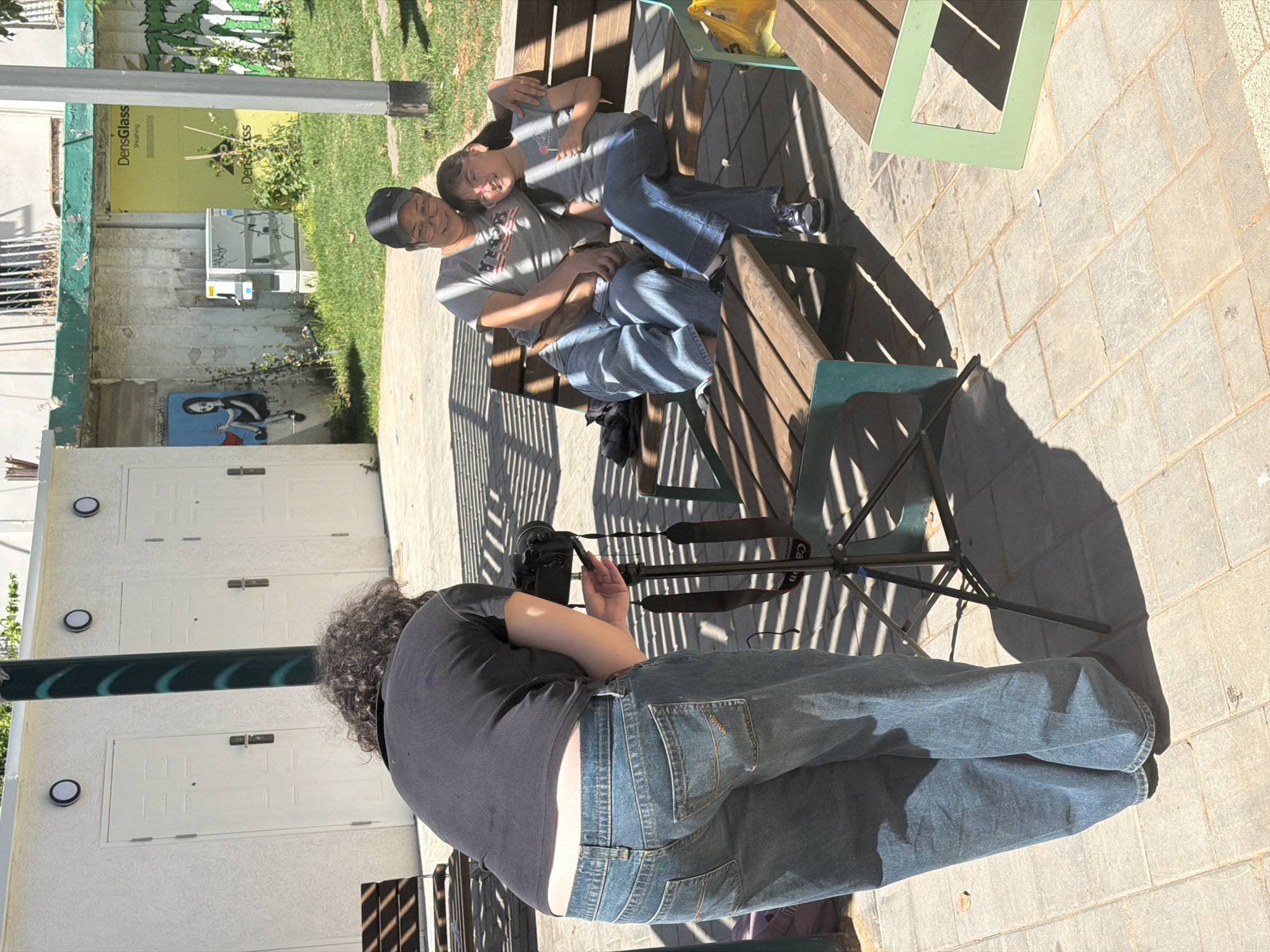 A person with curly hair is setting up a camera on a tripod to photograph a couple sitting on a wooden bench. The couple appears relaxed, with one person lying down on the lap of the other. The scene is outdoors, likely in a park or public space in Tel-Aviv, as evidenced by Hebrew text on the side of a building in the background. The backdrop includes green vegetation, painted murals on walls, and outdoor furniture. The area is paved with rectangular stones.