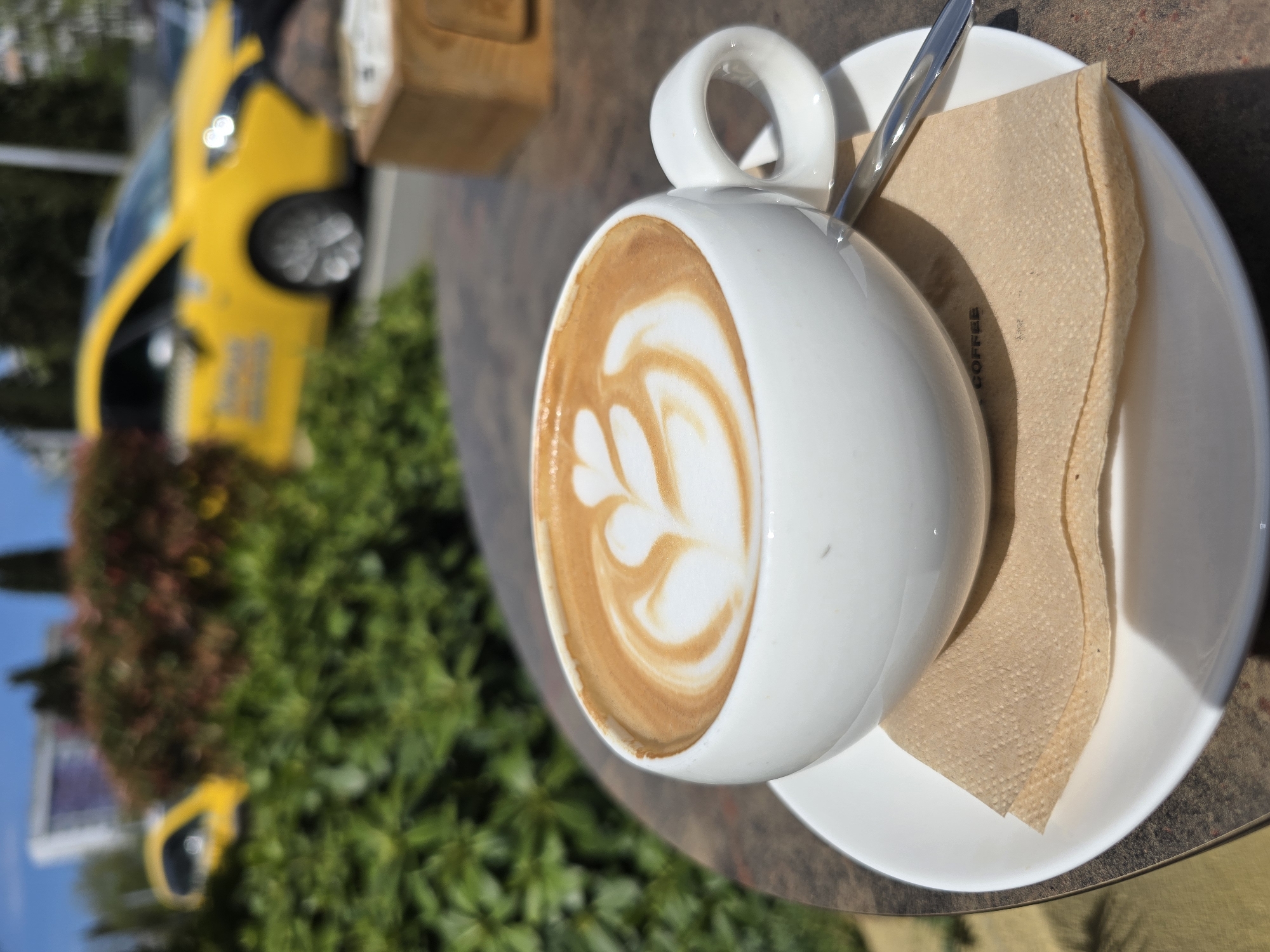 A white ceramic cup of coffee, featuring intricate latte art in a heart or leaf pattern, takes center stage on a white saucer. A silver spoon rests on the saucer, partially hidden by the cup, alongside a folded, light brown paper napkin with the word "COFFEE" faintly visible. The cup and saucer are positioned on a dark, textured tabletop, suggesting an outdoor cafe setting.
The background is intentionally blurred, directing focus to the coffee. Through the blur, a distinctive bright yellow vehicle, possibly a taxi, is visible on the left side, hinting at a nearby street or parking area. Green foliage, likely bushes or trees, forms part of the soft background, further indicating an outdoor environment. The scene is bathed in bright, natural sunlight, suggesting a clear day, possibly morning or early afternoon, and casting subtle shadows. The overall impression is one of leisure and enjoyment, capturing a relaxed moment outdoors in Burgas, Bulgaria.