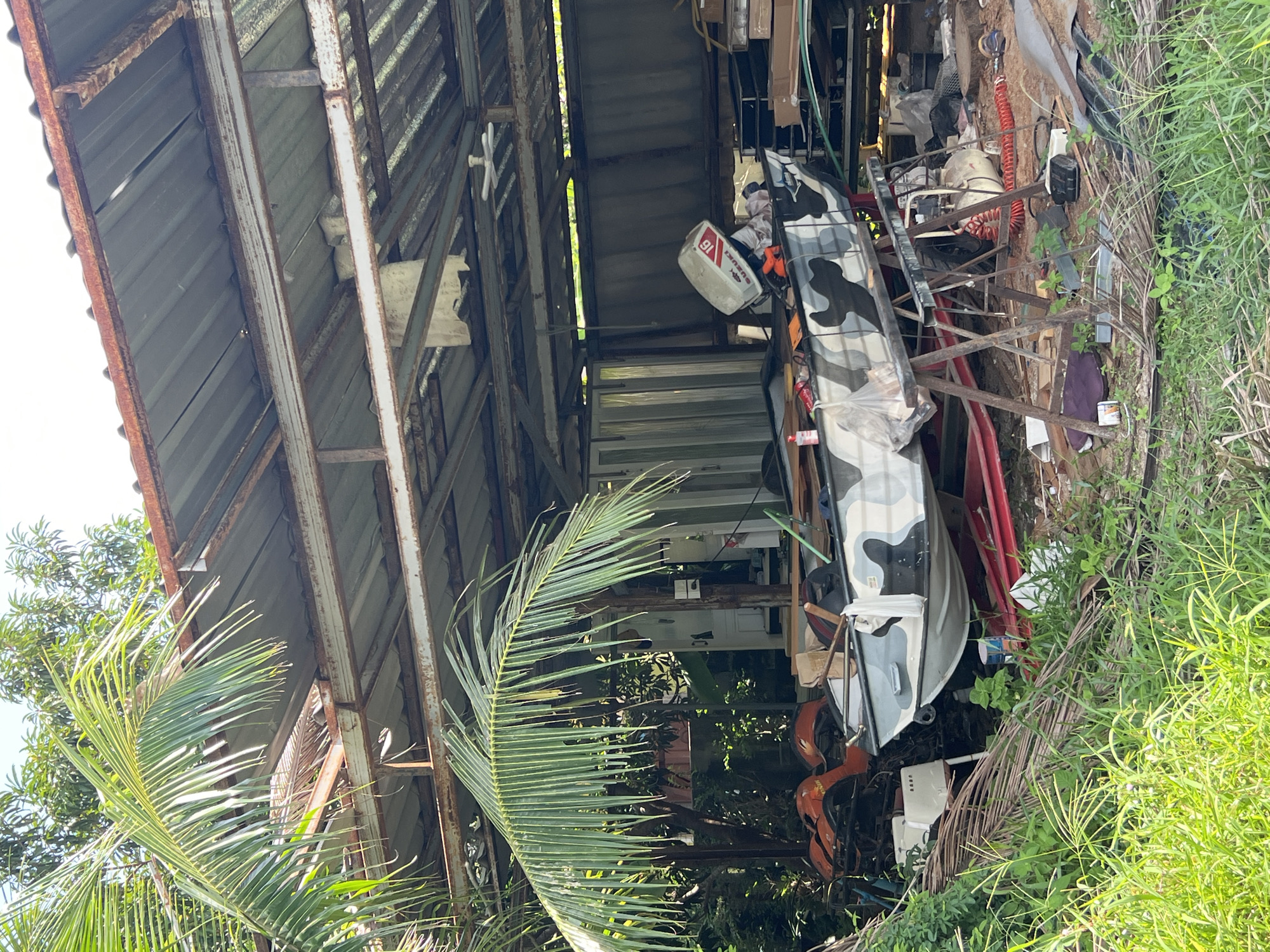 The image depicts a cluttered outdoor storage area, possibly a boat shed or workshop, located in Choeng Thale, Thailand. Dominating the foreground is a camouflage-patterned boat resting on a red stand with metal supports. The boat has a white and gray camouflage pattern, and a white outboard motor with red and black markings is mounted on its stern. To the left of the boat, part of an orange ATV is visible. Scattered around the boat and in the background are various items including wooden planks, a white cylindrical object that appears to be an air compressor with red coiled tubing, assorted tools, and debris. A large, green palm frond extends from the left side of the frame, partially obscuring a weathered metal roof structure supported by rusty metal beams. The structure appears to be open on at least one side, allowing natural light to filter in. In the background, more vegetation and what looks like corrugated metal fencing can be seen. The ground is covered in grass and dirt. There are no people visible in the scene.