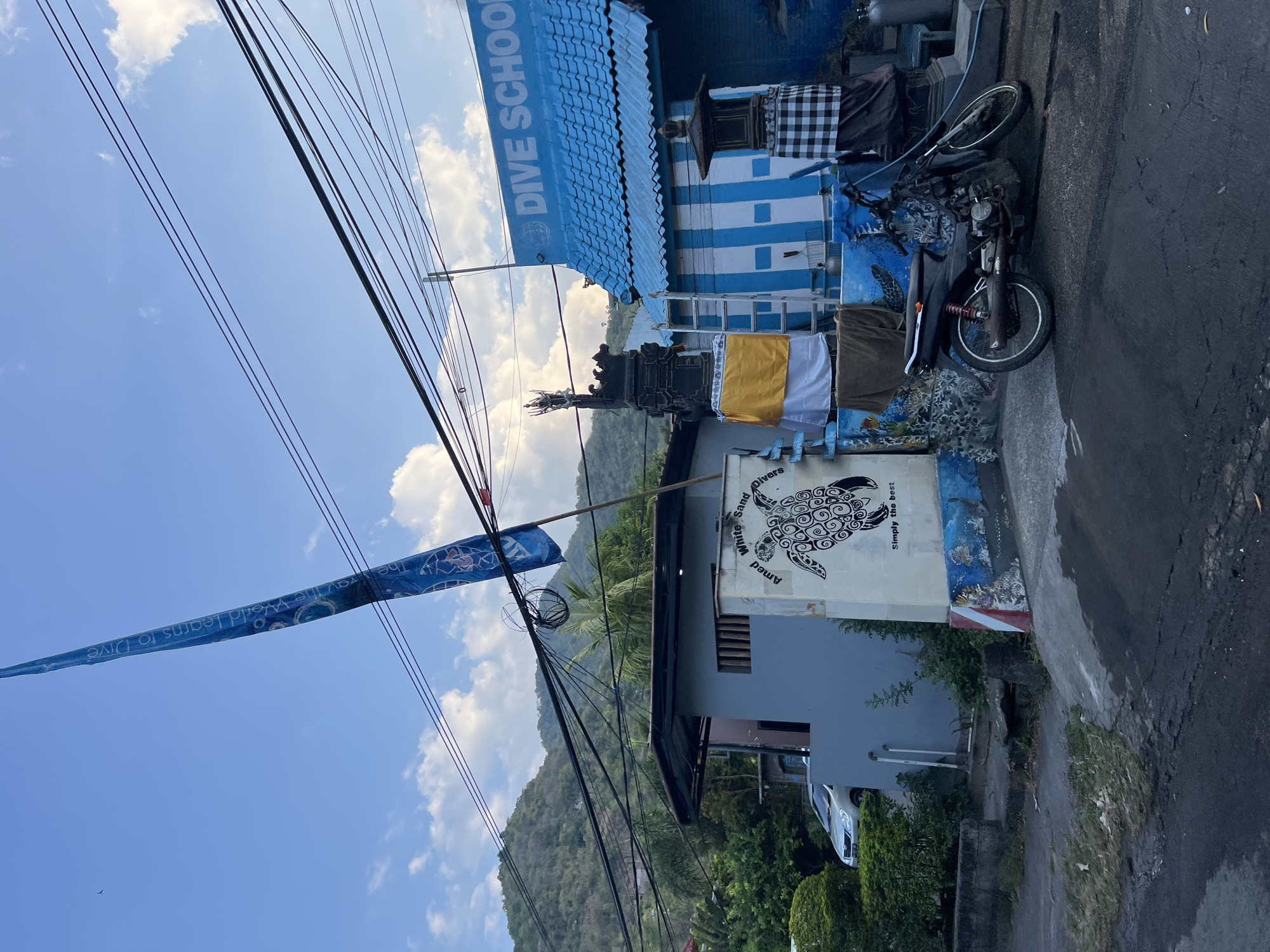 In the city of Abang, Indonesia, this photograph captures the facade of a dive school. A bright blue building with white striped walls and a blue corrugated roof is prominently displayed. Above the entrance, the words "DIVE SCHOOL" are written in white lettering. To the left of the building, a tall, narrow blue flag with white text and circular designs stands at an angle against a cloudy blue sky. A black motorcycle is parked in front of the dive school. Adjacent to the dive school, there's a gray building with a dark gray roof. A white wall with a mural of a sea turtle and the text "Amed White Sand Divers" and "Simply the best" is positioned between the gray building and the dive school. A black and white checkered cloth hangs on a structure to the right of the dive school. In the background, lush green trees and a mountainous landscape are visible. Overhead, a dense network of black electrical wires crisscrosses the scene.