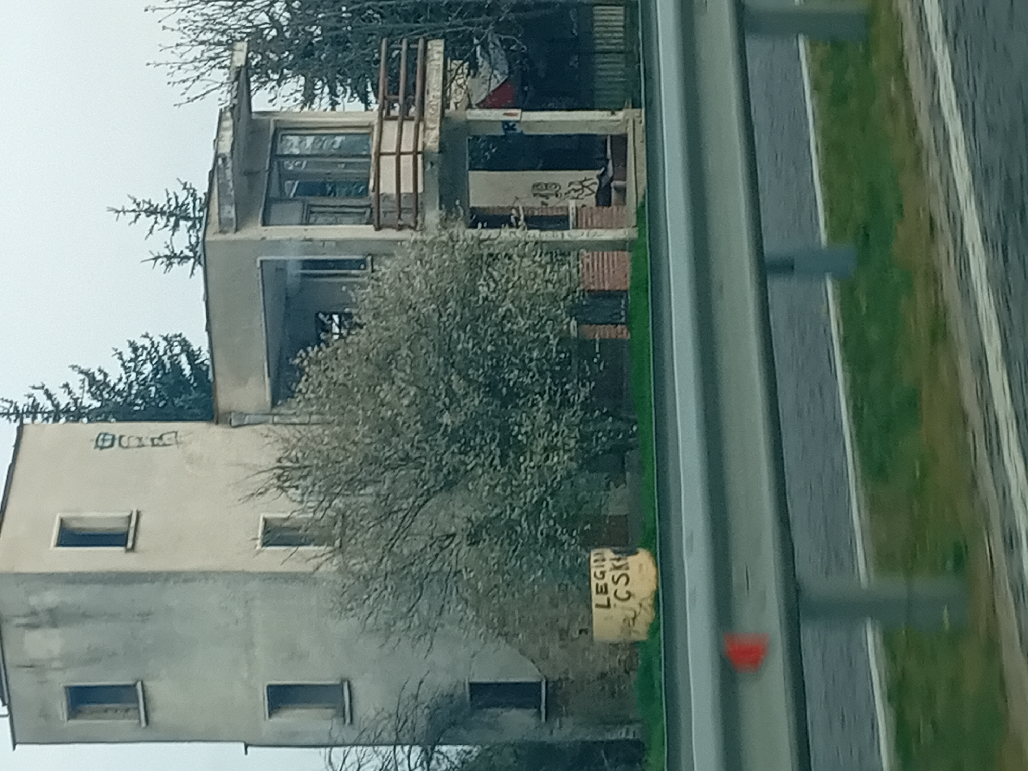 The image shows a tall, dilapidated building located near a road with a guardrail. The building appears to be multi-story, with missing windows and visible signs of disrepair. Graffiti is visible on the upper floors. A tree partially obscures the building. A sign is present near the base of the building, with the text "LEGII CSK" visible. The surrounding environment includes grass and trees alongside the road. The location is described as "Unknown, Unknown".