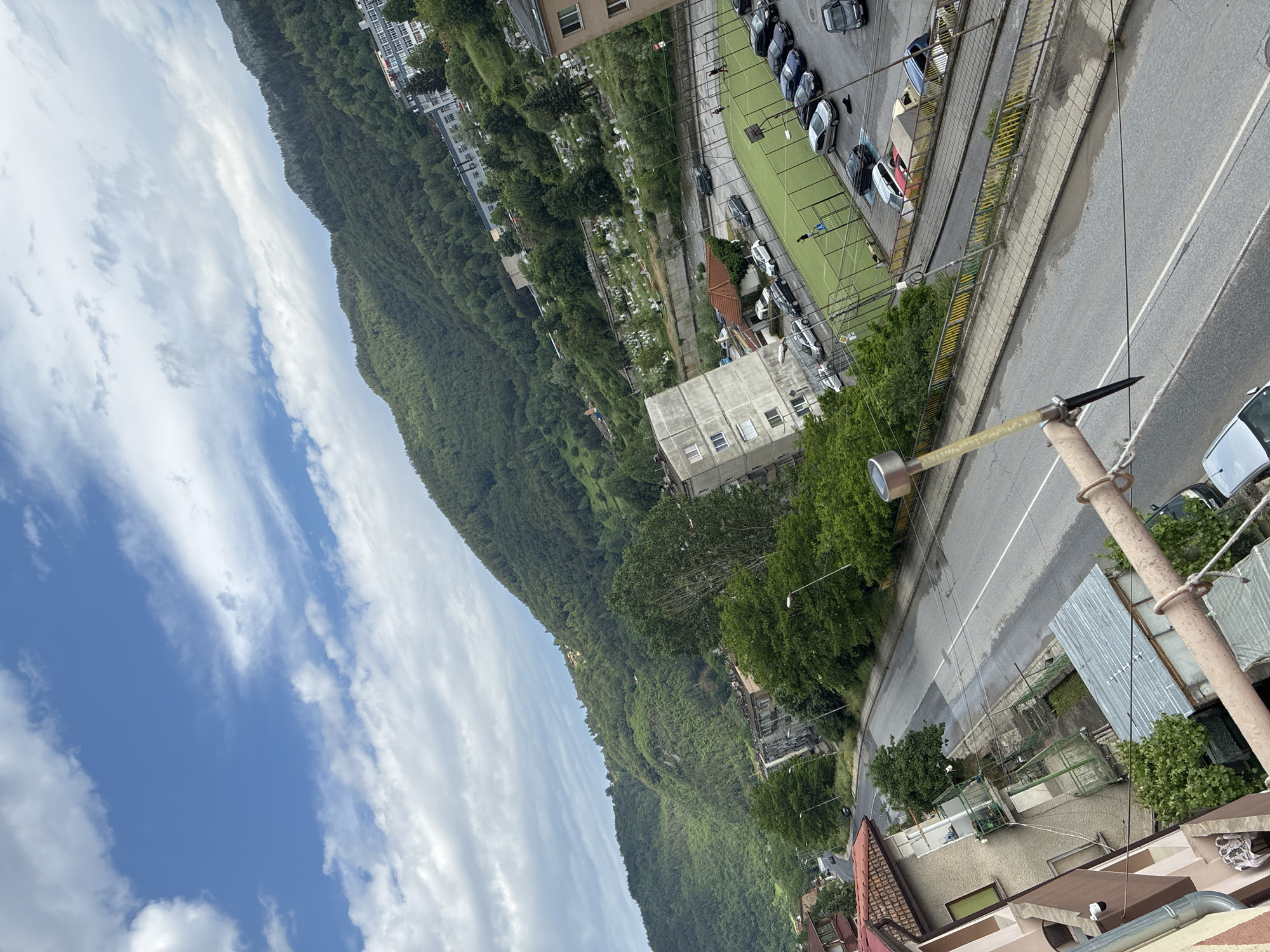 The image presents an elevated, diagonally tilted view of a street and surrounding urban and natural landscape, located in Vlahovo, Bulgaria. The sky occupies the upper portion, featuring a light blue expanse with scattered cumulus clouds of varying density.
In the background, densely forested mountains rise, covered predominantly by green trees. On the slopes of these mountains, several small residential structures with light-colored facades are visible, blending with the natural environment.
The midground features a transition between dense vegetation and urban structures. Towards the upper right, on a gentler slope, a cemetery is visible, characterized by numerous white or light-colored gravestones arranged closely amidst green foliage. Below the cemetery and to its left stands a prominent multi-story building with a light-grey, somewhat weathered facade, featuring multiple windows and balconies. To the right of the cemetery, another multi-story building with a lighter facade and numerous windows is partially visible.
In the central midground, a green artificial turf multi-sport court, marked with white lines and equipped with goals at both ends, is present. It is enclosed by a dark fence. Approximately three to four individuals are discernible on the field, appearing to be engaged in activity or walking. Surrounding the sport court and along the adjacent road, numerous cars of various makes and colors (including silver, black, dark blue, red, and white) are parked.
A two-lane asphalt road runs diagonally from the lower-left to the mid-right of the image. It has a visible white center line and some faded white shoulder markings. The road surface appears somewhat damp or uneven in sections. Parallel to the road are sidewalks, delineated by curbs and, on the right side, by metal railings painted in yellow and green. Streetlights are positioned along the road.
On the immediate left side of the foreground, part of a multi-story building with a brown and beige facade and a red-tiled roof is visible. This building features balconies with green railings, and a security camera is mounted on its exterior. Below it, a smaller structure with a corrugated metal roof is partially visible.
In the immediate foreground, a metal railing, appearing aged and partially painted brown, spans horizontally, likely part of a balcony. A solar-powered garden light with a transparent, yellowish stem and a dark, spiked base is attached to this railing. Several black wires run horizontally across the lower frame of the image.