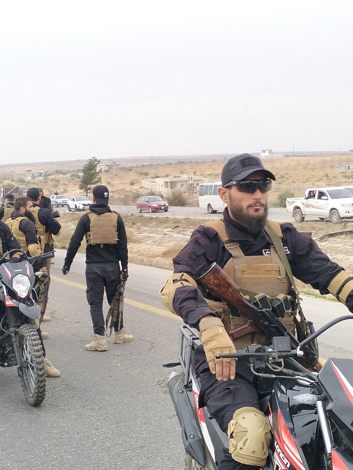 A group of armed men, possibly security or military personnel, are lined up on a road near a desert landscape with sparse buildings. They wear dark clothing, tactical vests, and caps, some with sunglasses. Several are on motorcycles, and others stand holding rifles. Cars and a white bus are visible on the road in the background. The overall tone suggests a security presence or operation.