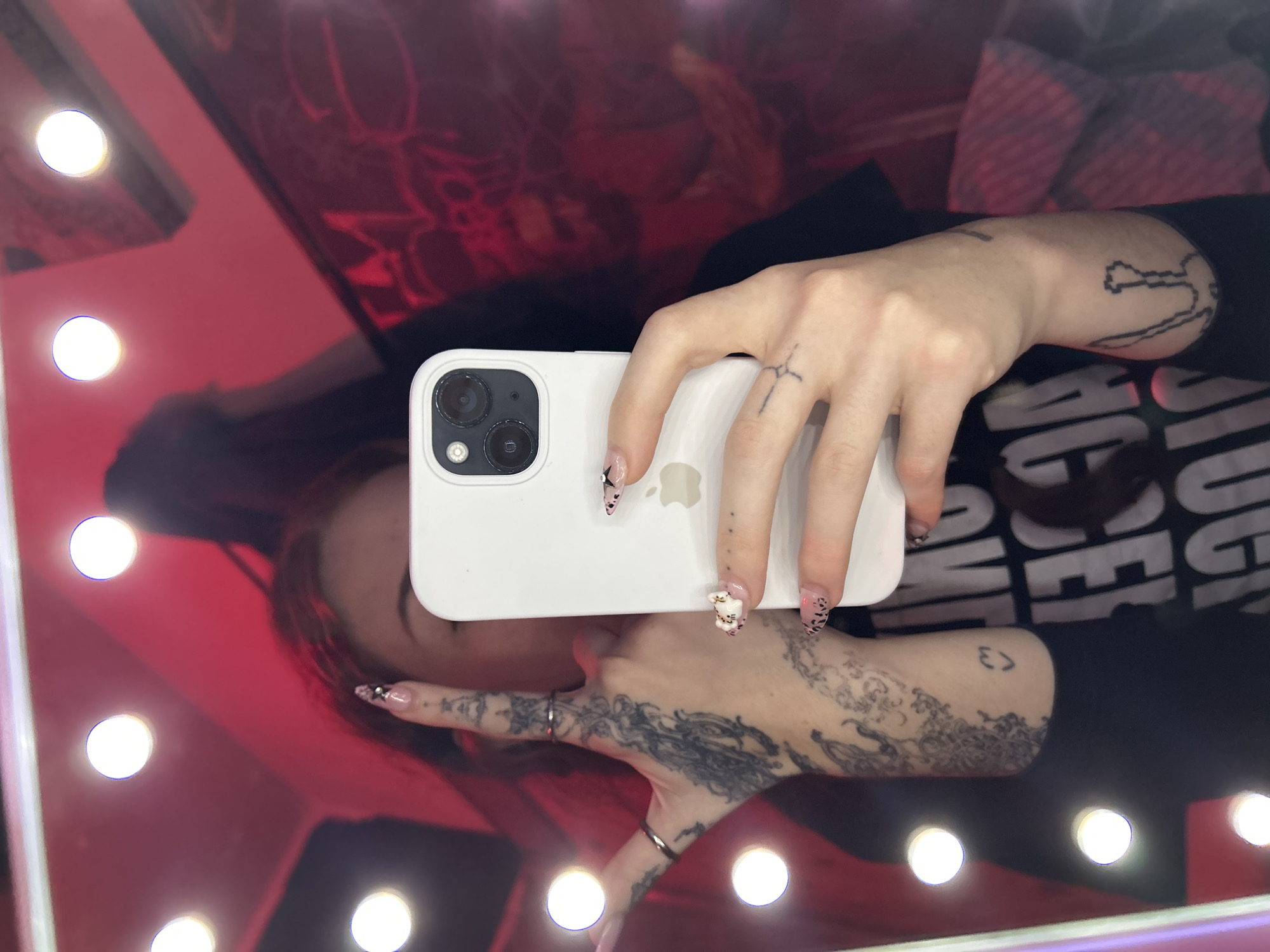 A person with tattoos is taking a selfie in a mirror with an iPhone. The mirror is surrounded by lights, and the background is a blurry red color. The person is wearing a black shirt. It is not possible to tell the location.