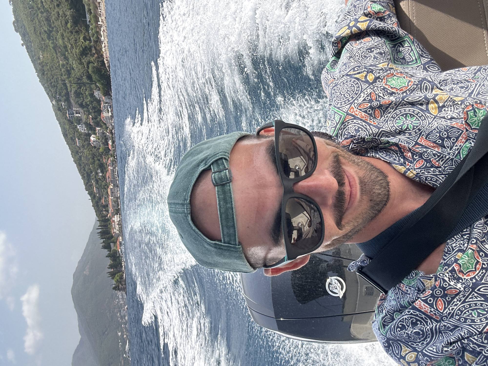 A man wearing sunglasses and a green baseball cap is taking a selfie on a boat. The boat is moving, creating a wake in the blue water. In the background, there is a coastline with hills covered in trees and houses. The scene appears to be the Bay of Kotor in Montenegro, specifically near the Luštica peninsula. The man is wearing a colorful, patterned shirt and a black strap across his chest. The sun is shining, and the sky is blue with some clouds.